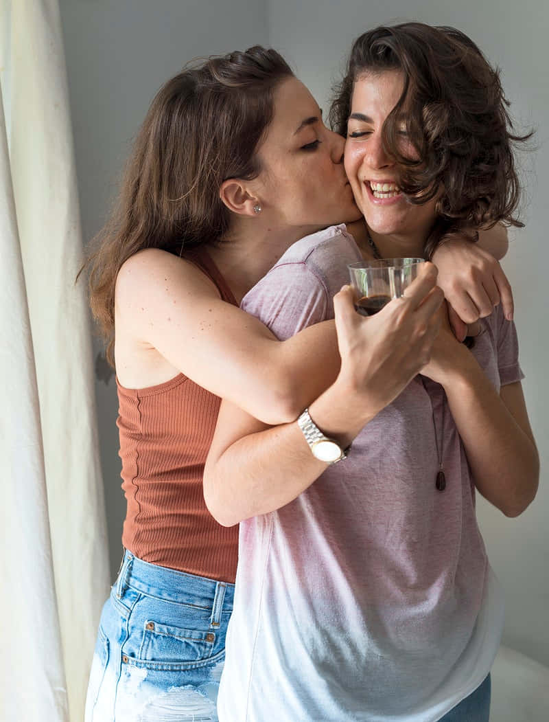 Happy Couples 桌布 壁紙 — 兩位女士在室內熱情擁抱，其中一位笑著親吻另一位臉頰，同時拿著咖啡杯。