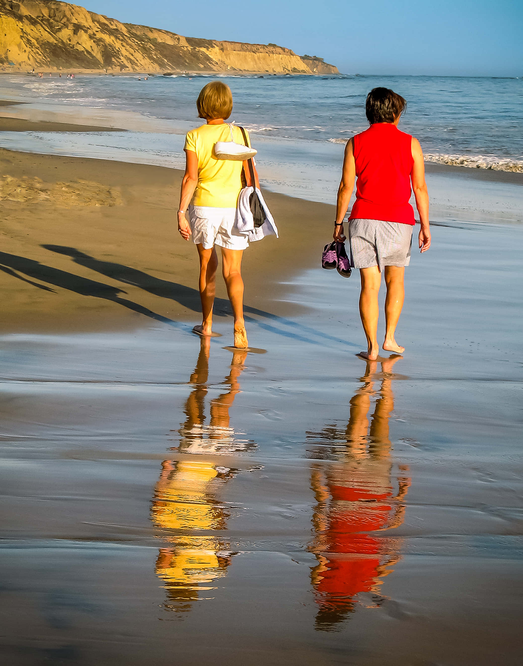 Beach Walk 桌布 壁紙 — 兩位女士在日落時赤腳漫步於濕沙灘上，她們的倒影在淺水中閃爍。