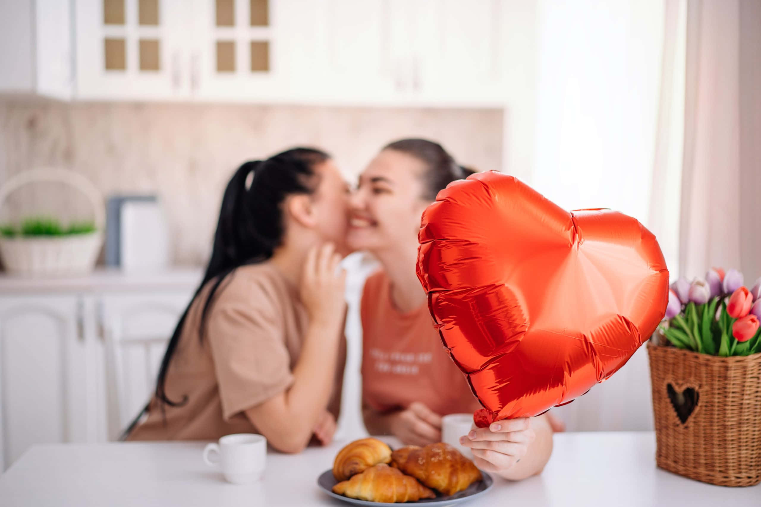 愛心 桌布 壁紙 — 兩位女士在明亮的廚房裡分享一個快樂的吻，同時拿著一個紅色心形氣球和可頌。