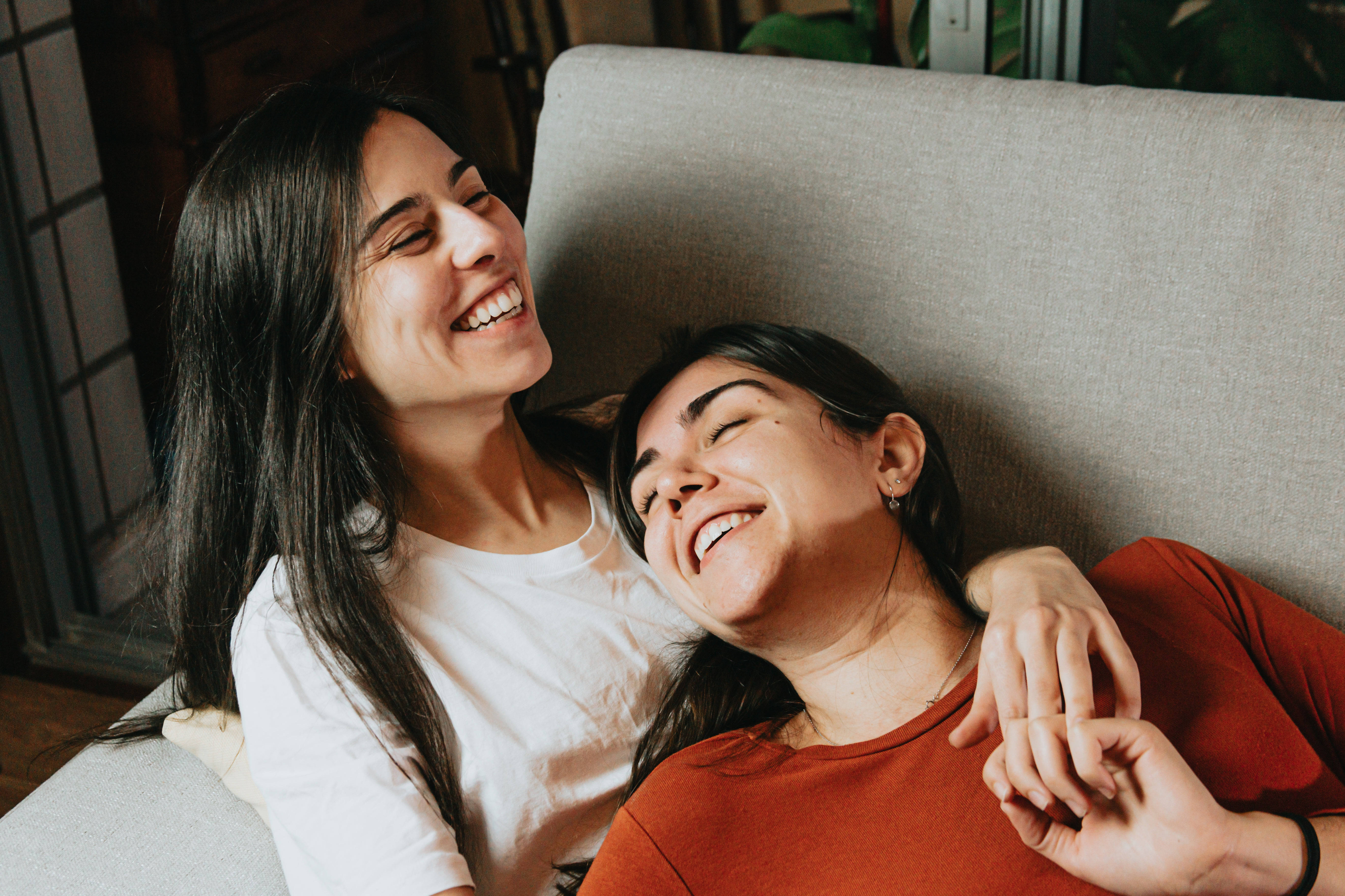 Love And Friendship 桌布 壁紙 — 兩位女士在溫暖燈光的室內灰色沙發上開懷大笑並擁抱。