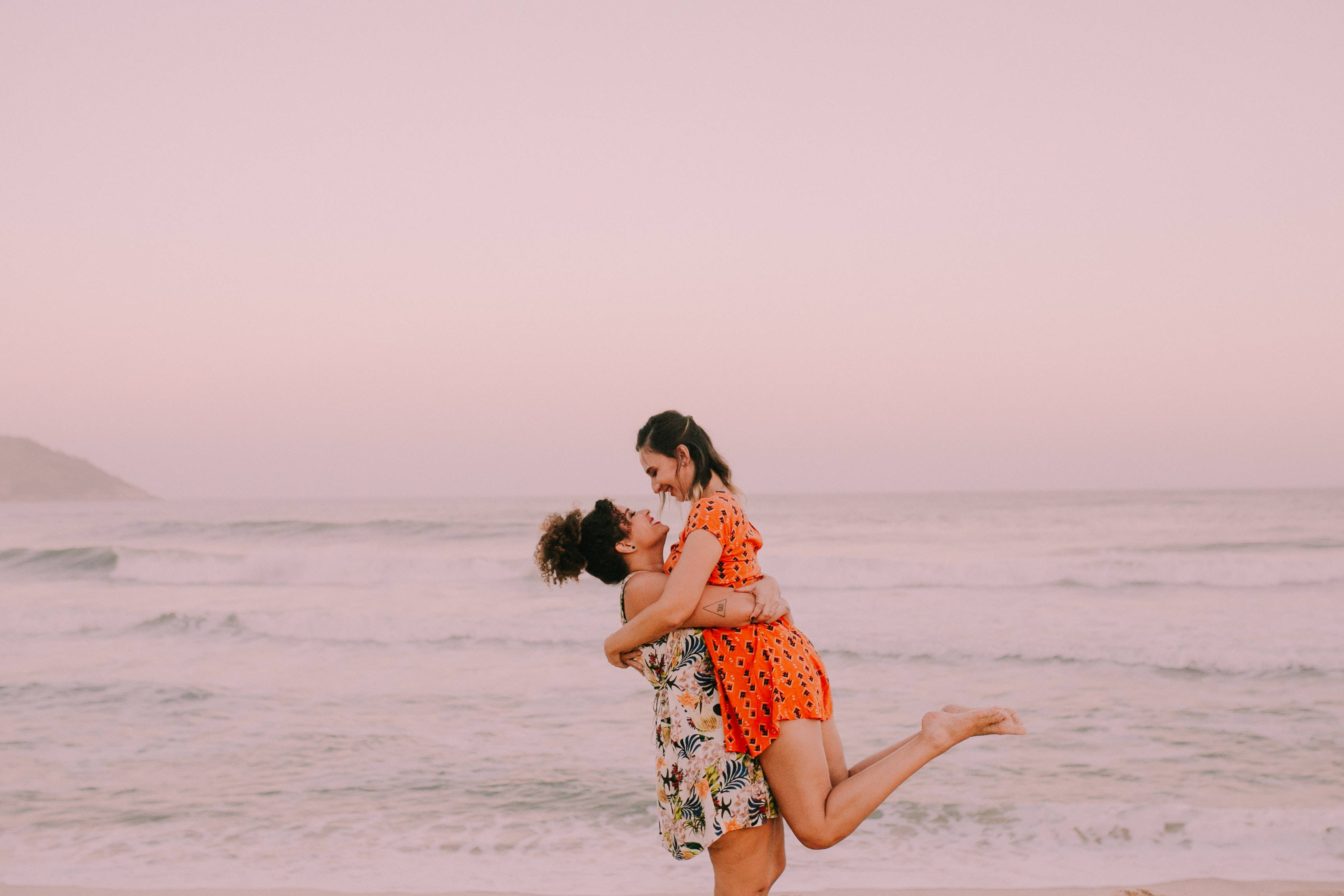 couple love 桌布 壁紙 — 兩位女士在粉紅色海灘上日落時分愉快地擁抱，伴隨著溫柔的海浪和遠山。
