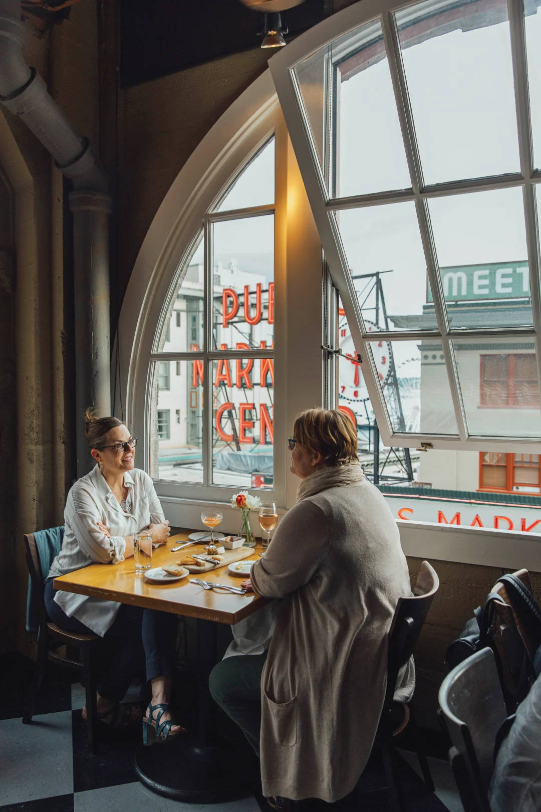 Pike Place Market 桌布 壁紙 — 兩位女士在舒適的咖啡館裡，在一張大拱形窗戶下的木桌上享用飲品並聊天。