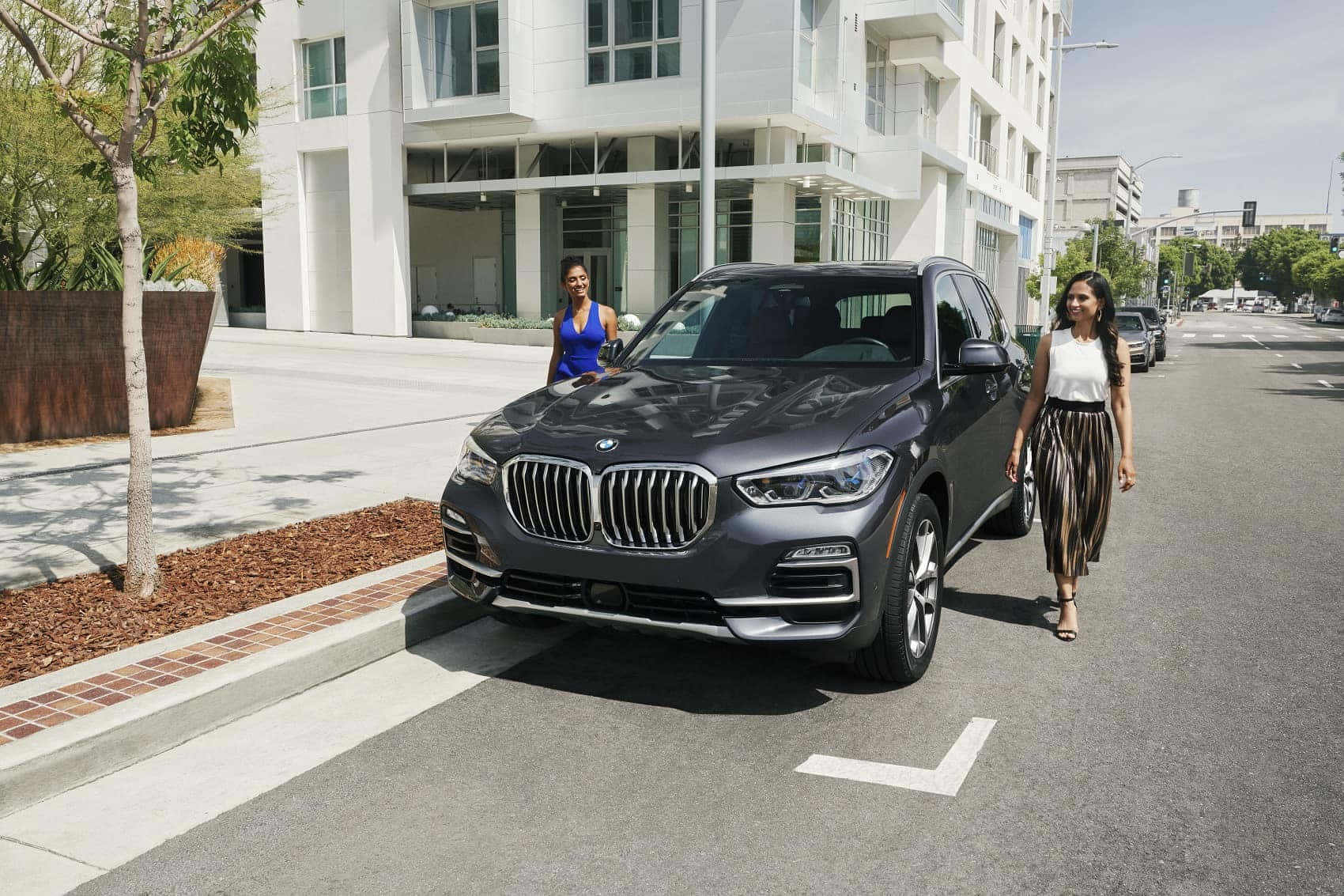Bmw X5 桌布 壁紙 — 兩位女士在陽光明媚的城市街道上，在一輛停放的時尚灰色BMW SUV旁邊散步，周圍有現代建築和樹木。