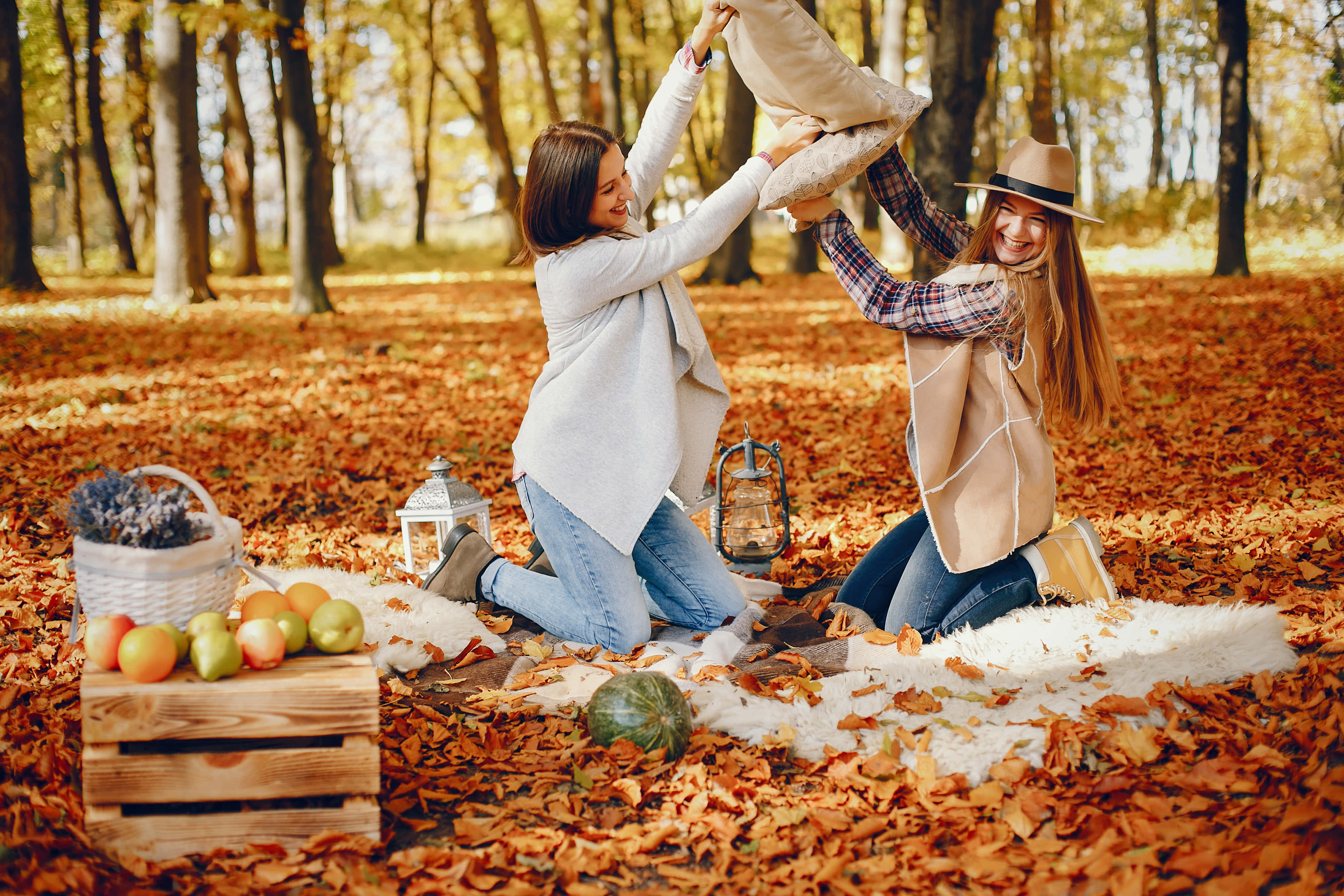 Fall Family 桌布 壁紙 — 兩位女士在陽光明媚的秋日森林中歡快地扔枕頭，周圍是落葉和野餐佈置。