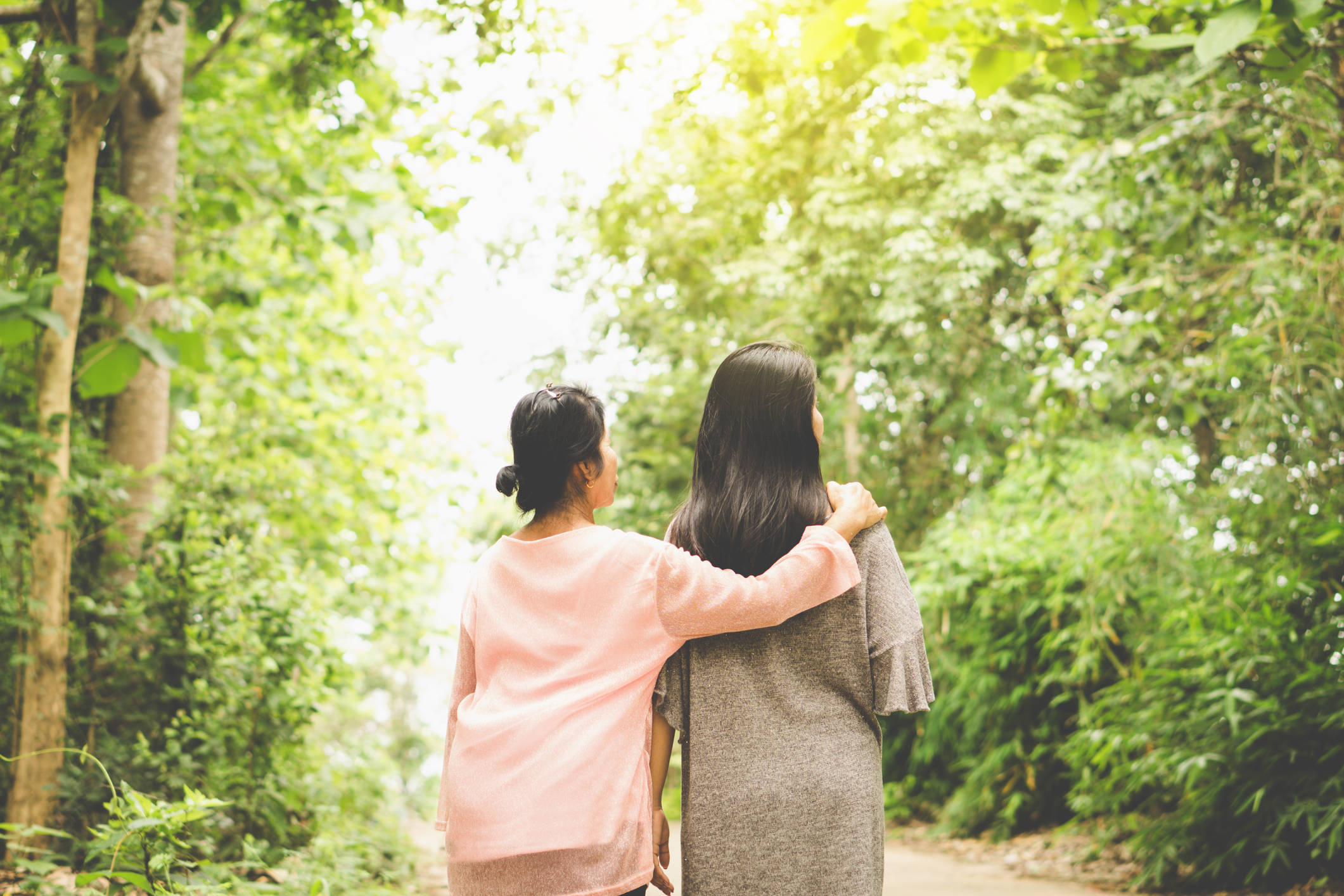 森林 桌布 壁紙 — 兩位女士手挽手走在一條陽光斑駁的森林小徑上，周圍是茂密的綠色樹葉。