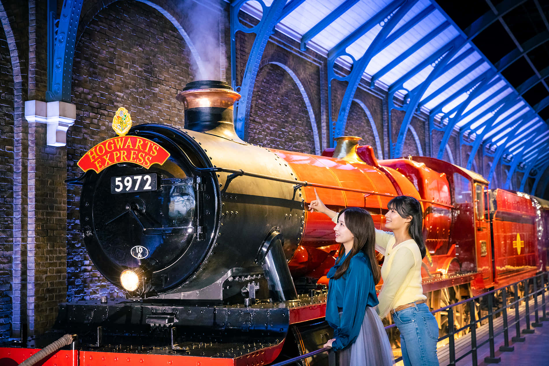 Hogwarts Express 桌布 壁紙 — 兩位女士欣賞在藍色拱形車站燈光下閃耀的標誌性霍格華茲特快蒸汽火車。