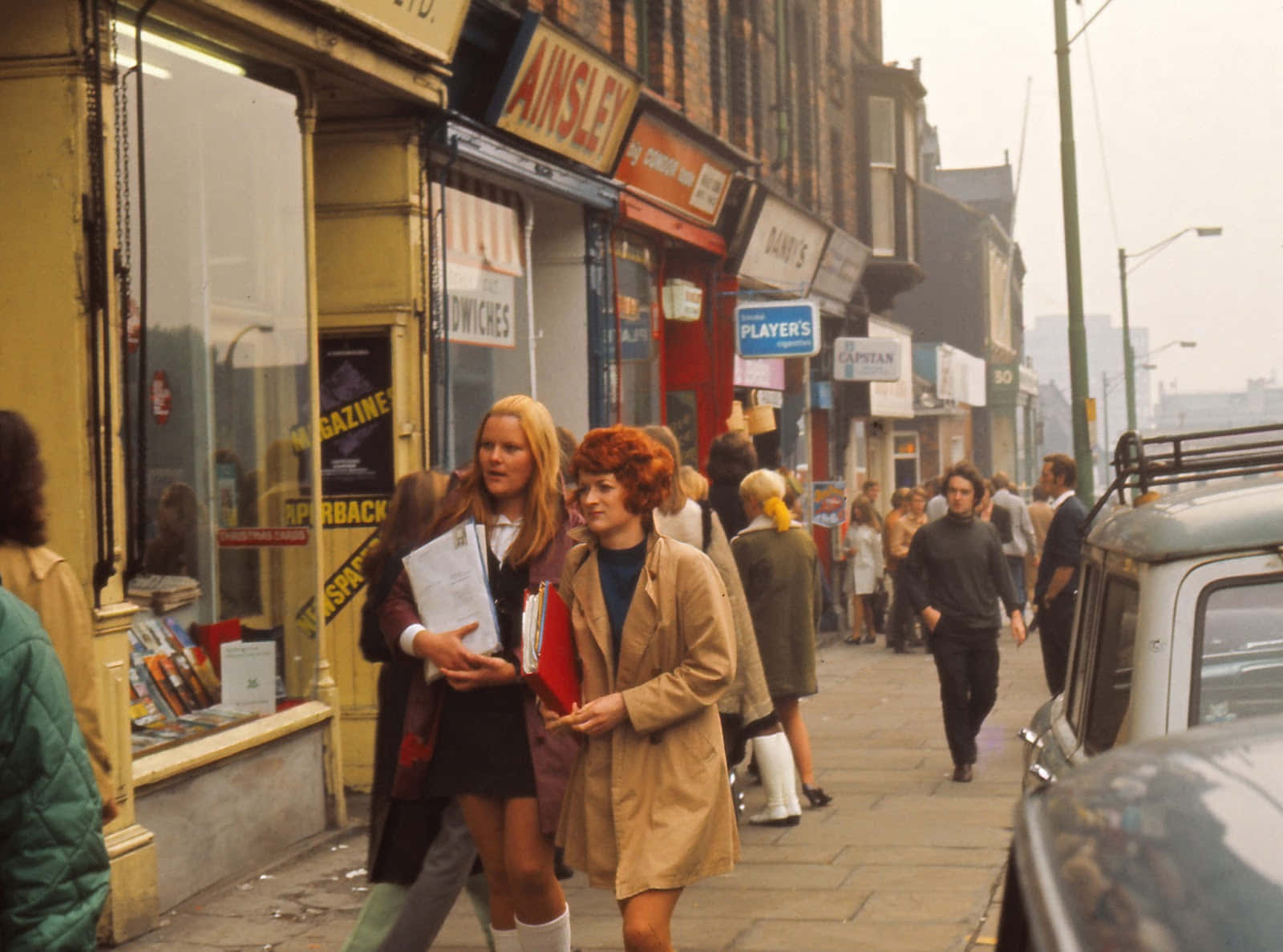 1970s 桌布 壁紙 — 兩位女士沿著1970年代英國繁忙的主要街道行走，經過商店和行人，路邊停著復古汽車。