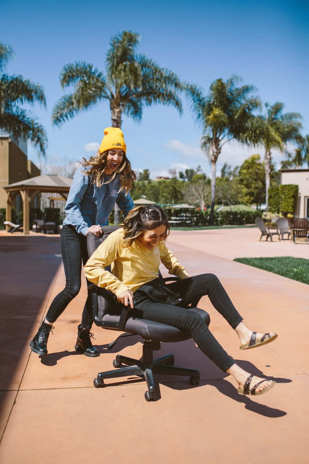 Bestie 桌布 壁紙 — 兩位女士笑著，其中一位在陽光普照、種有棕櫚樹的庭院裡推著另一位坐在辦公椅上。