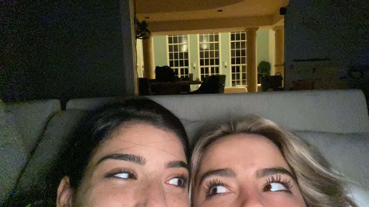 Charli And Addison 桌布 壁紙 — 兩位女性並排躺在沙發上，在昏暗的室內環境中帶著俏皮的表情向上看。