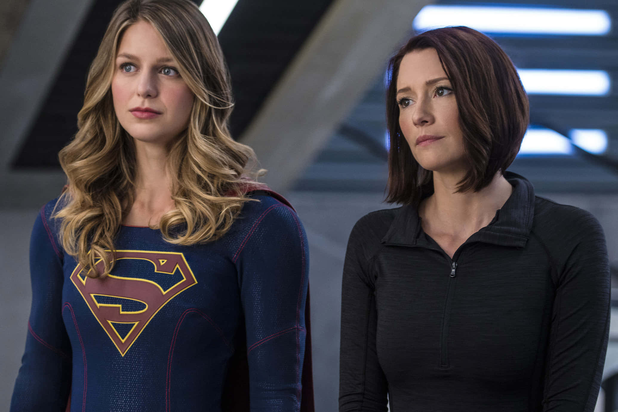Chyler Leigh 桌布 壁紙 — 兩位女性並肩站立，一位身穿藍色超人服，另一位身穿黑色拉鍊上衣，置身於未來金屬場景中。