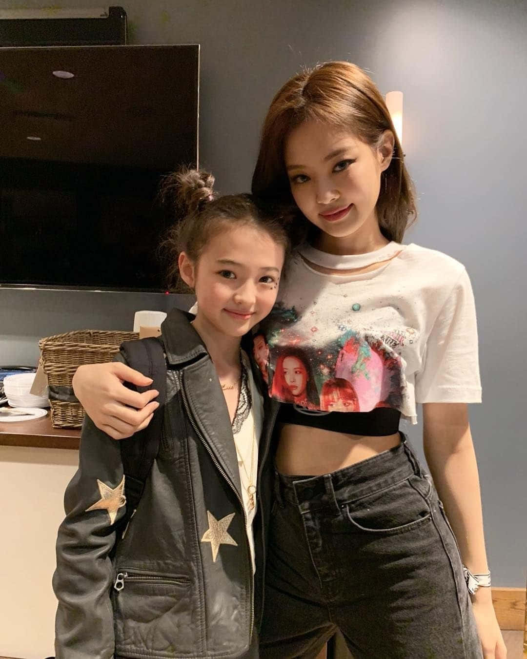 BLACKPINK 桌布 壁紙 — 兩位女性在室內溫馨地擺姿勢，一位身穿星星外套，另一位身穿圖案T恤，背景有電視和柳條籃。