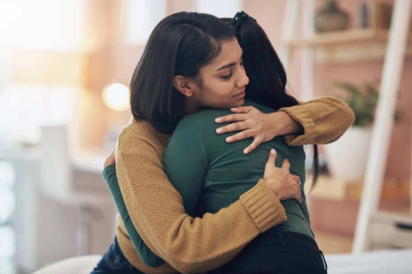 Hug 桌布 壁紙 — 兩位女性在柔和燈光的舒適室內空間中溫暖擁抱，背景有模糊的架子和植物。
