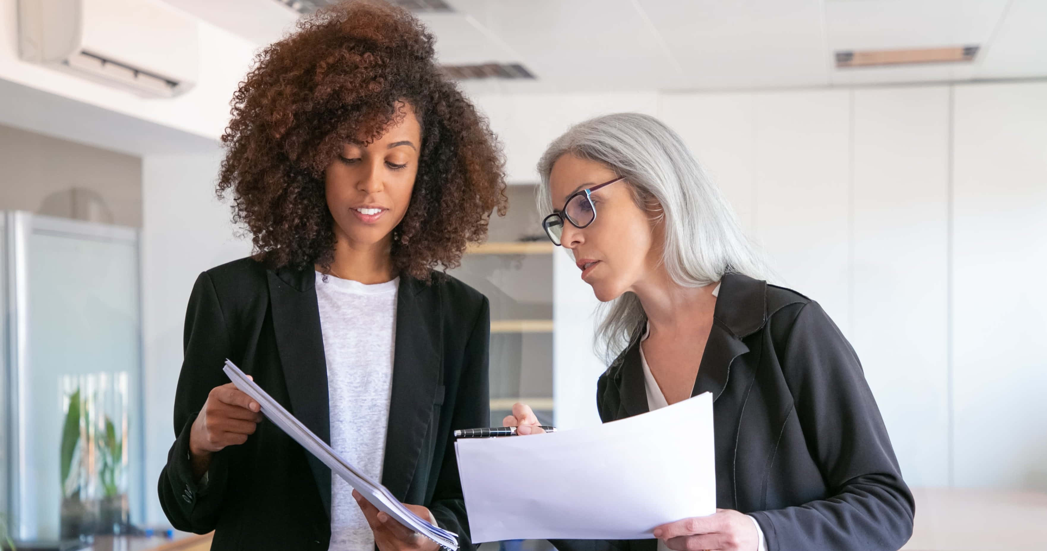 Leadership 桌布 壁紙 — 兩位女性在現代辦公室中，在明亮的頭頂燈光下，專注地審閱文件。