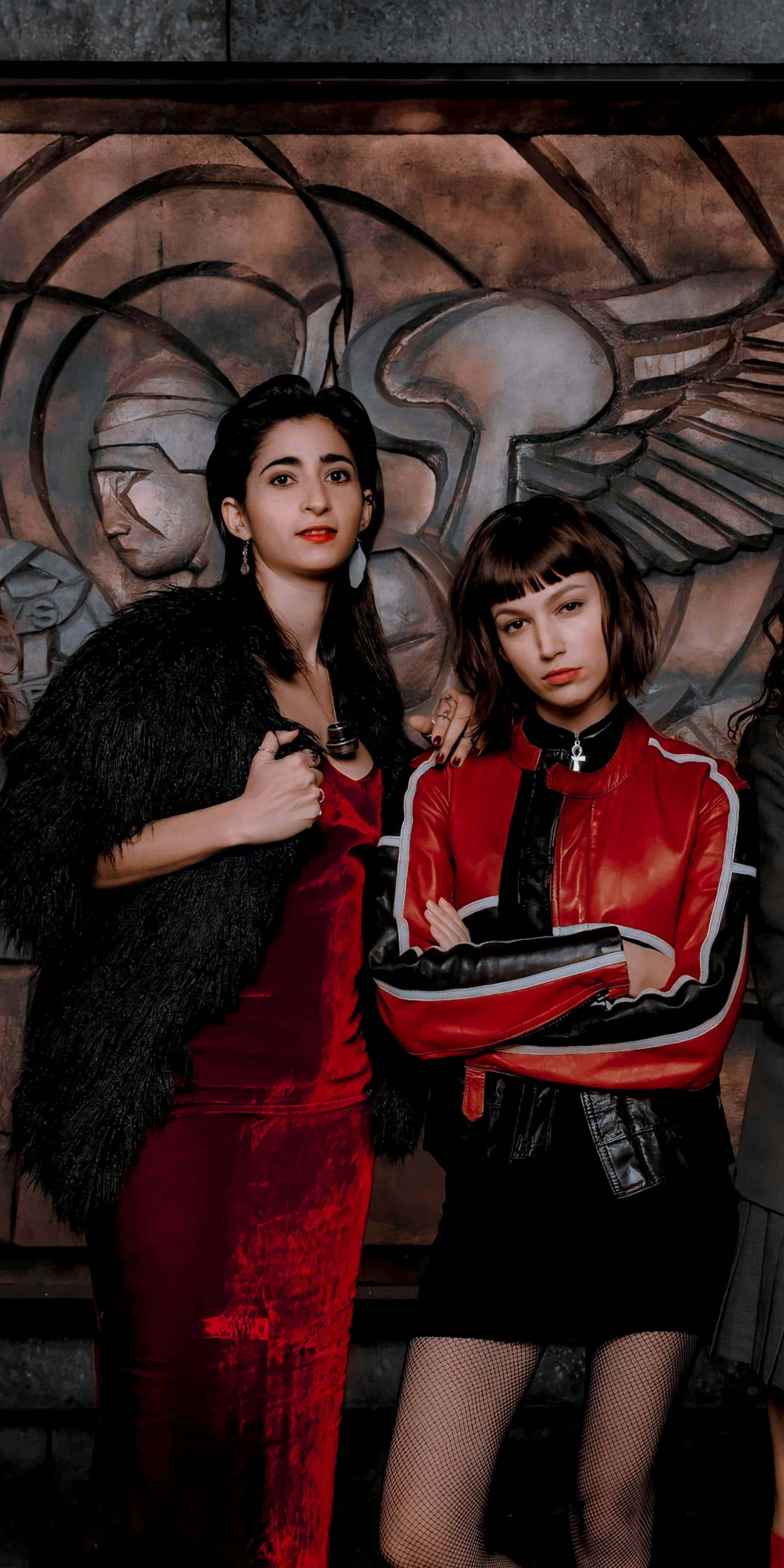 Money Heist Mask 桌布 壁紙 — 兩位女性在紋理壁畫背景前自信地擺姿勢，一位穿著紅色天鵝絨連衣裙配黑色毛皮披肩，另一位穿著紅色皮夾克配漁網襪。
