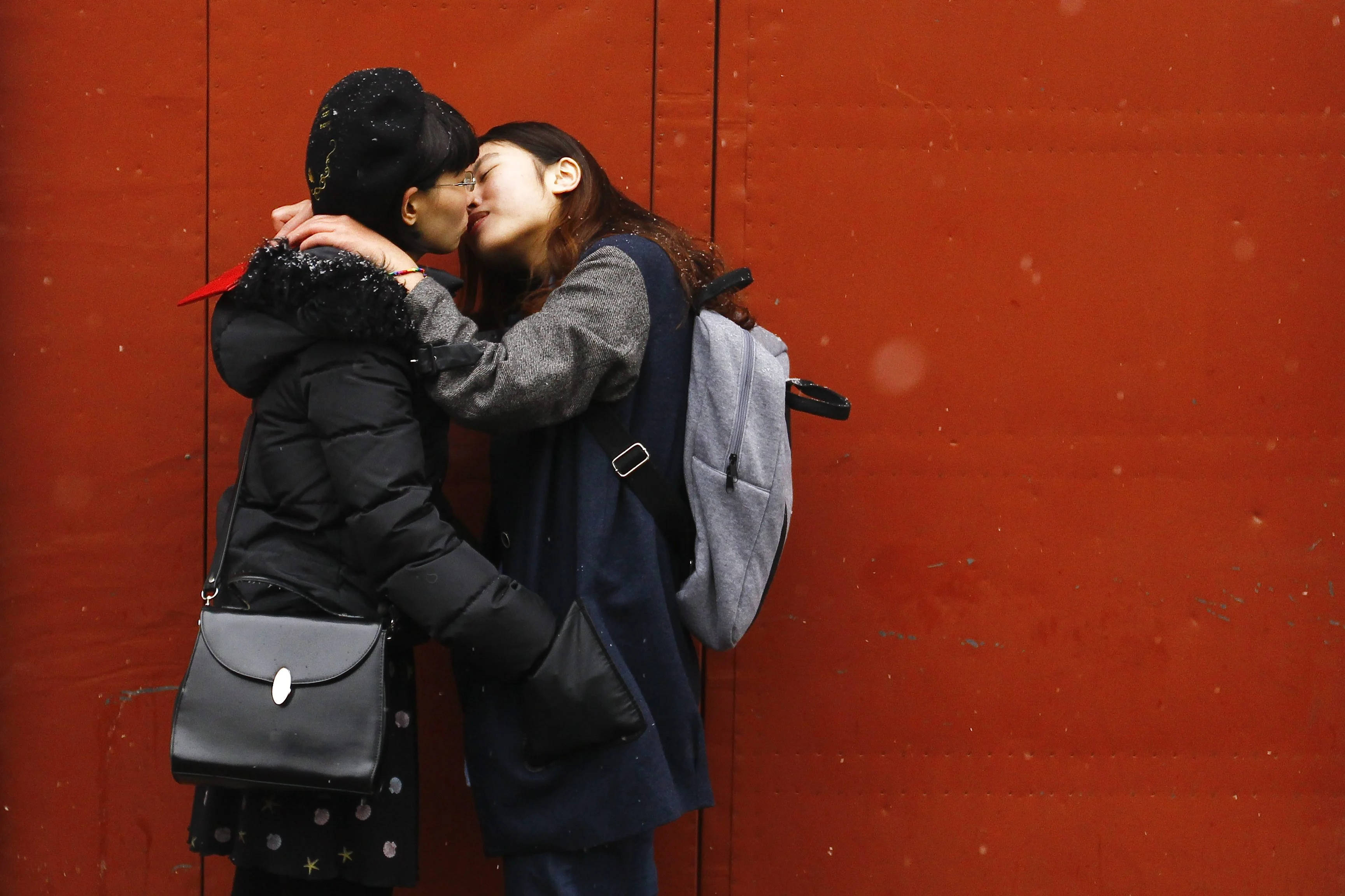 lesbian makeout 桌布 壁紙 — 兩位女性在鮮紅色的牆前熱情擁抱親吻，雪花輕輕飄落在她們周圍。