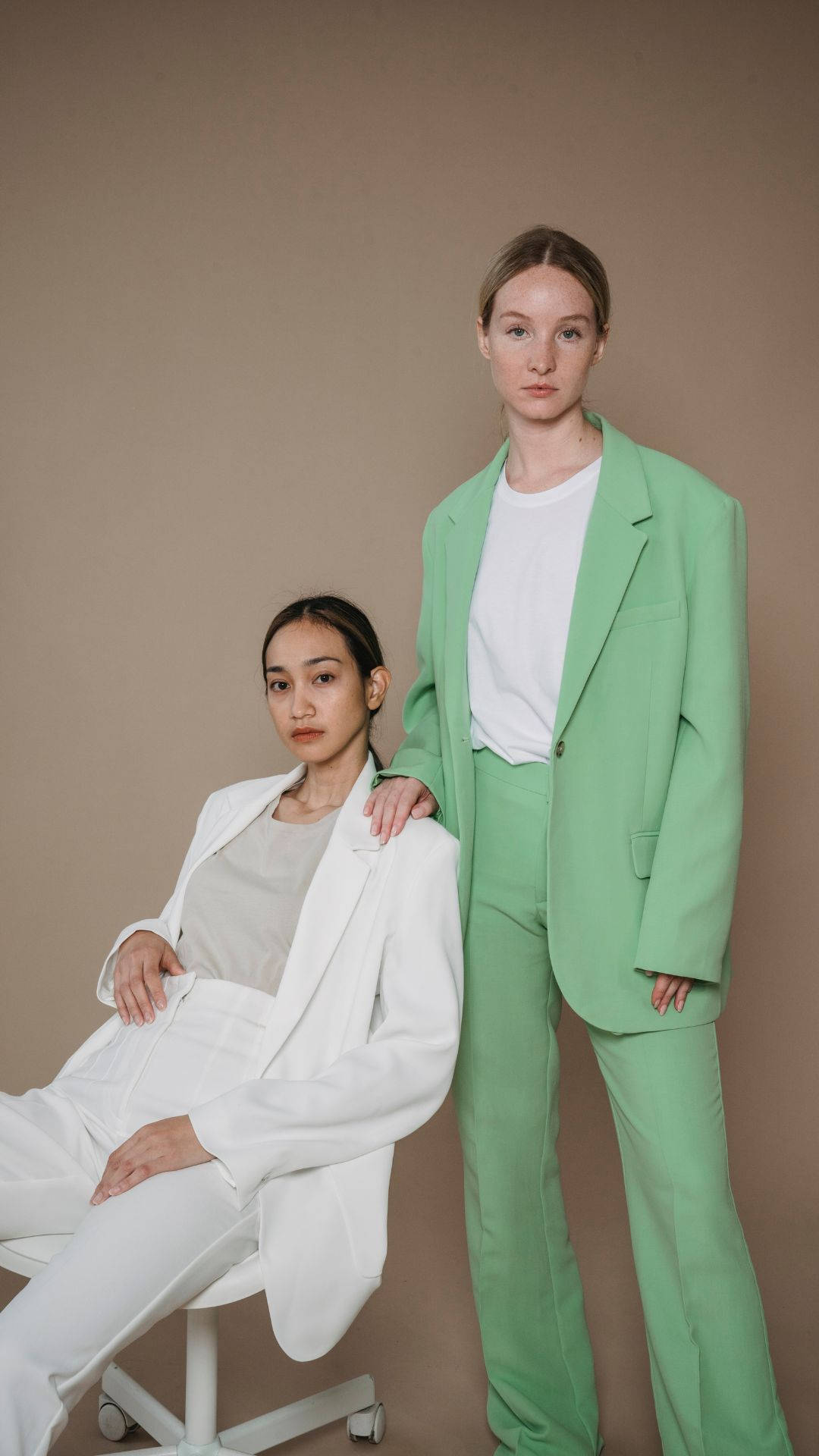 suit 桌布 壁紙 — 兩位女性自信地擺姿勢，一位身穿白色西裝坐著，另一位身穿薄荷綠色西裝站著，背景為中性米色。