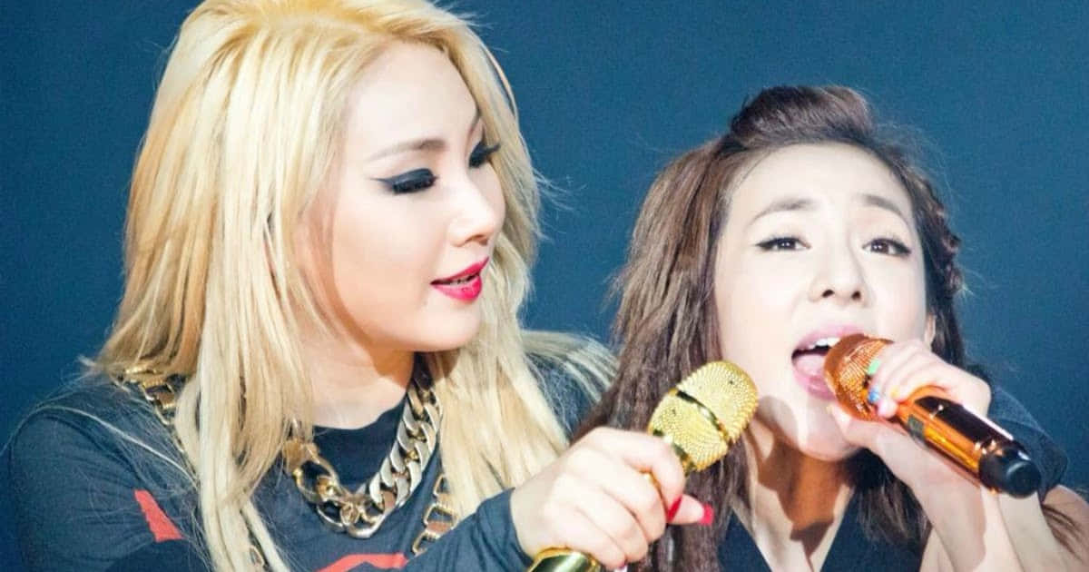 2NE1 桌布 壁紙 — 兩位女歌手在舞台上，在深藍色背景下，在明亮的舞台燈光下拿著麥克風表演。
