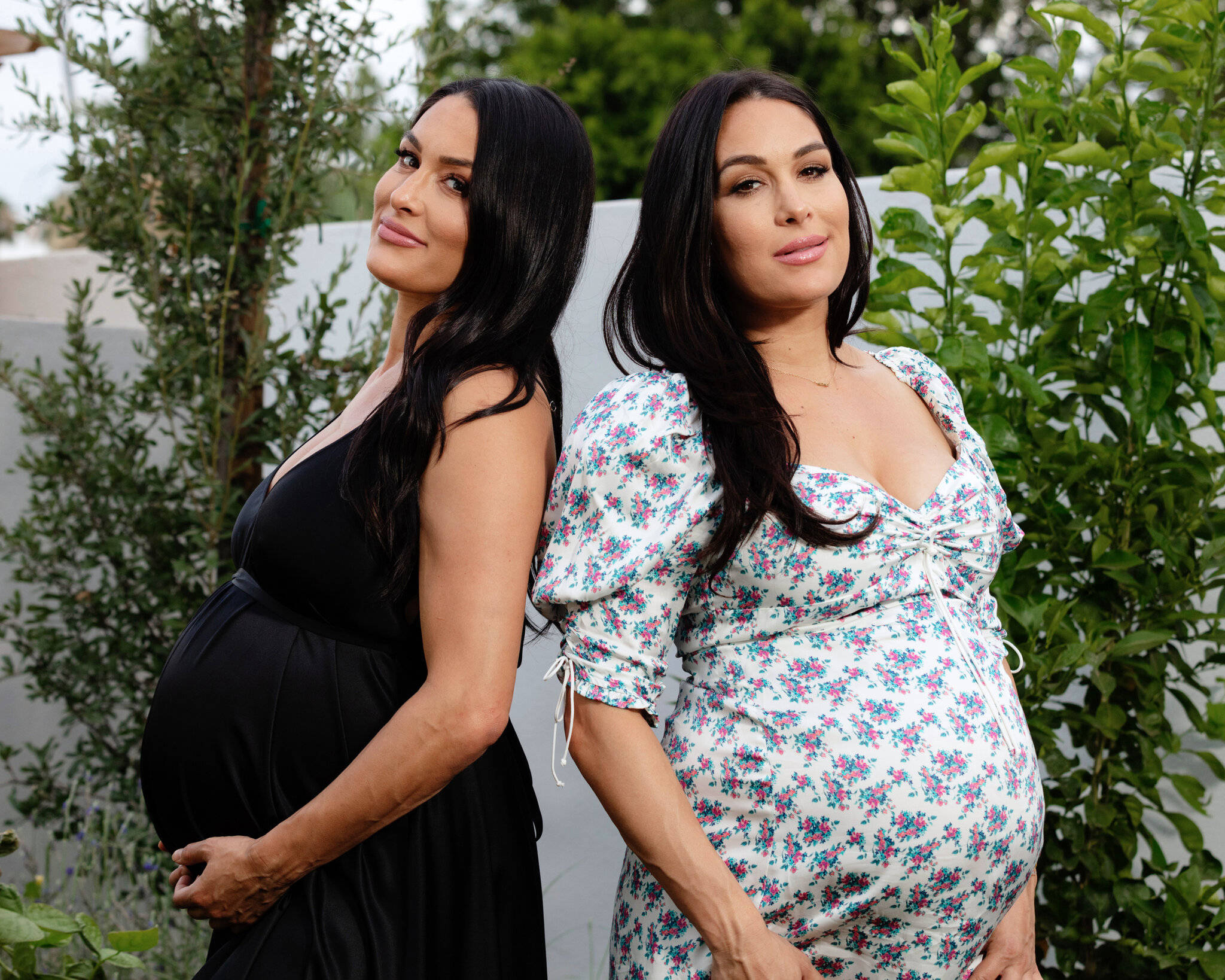 Pregnant 桌布 壁紙 — 兩位孕婦在花園中背靠背站立，穿著對比鮮明的連衣裙，一件黑色，一件花卉圖案，兩人都溫柔地微笑。