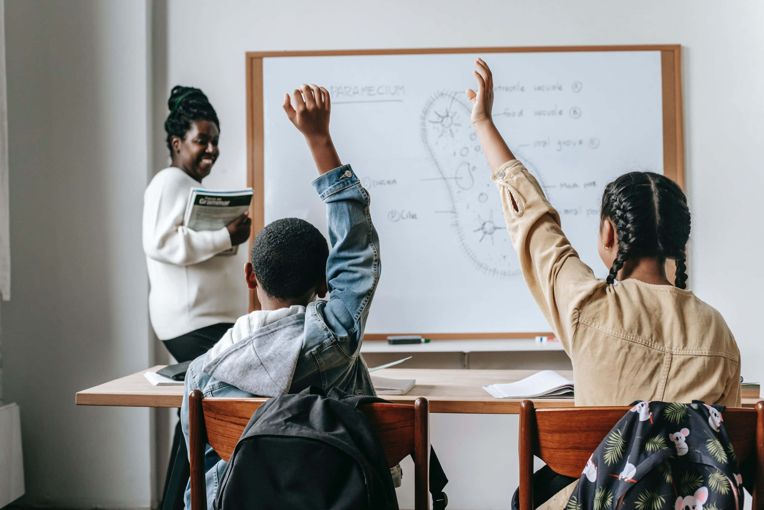 Teacher 桌布 壁紙 — 兩位學生在教室內熱情舉手，一位微笑的老師拿著書本，靠近帶有圖表的白板。
