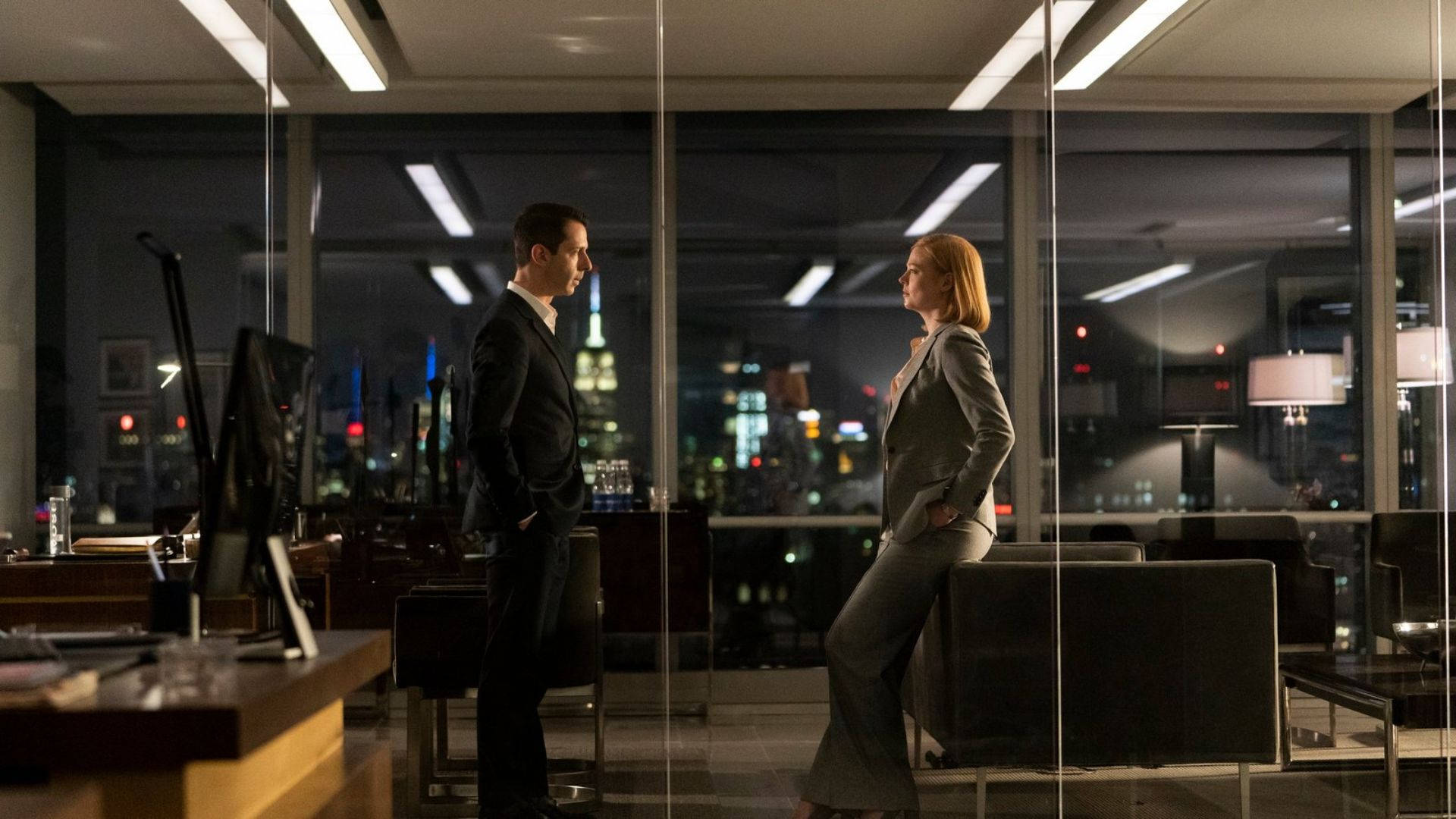 Succession 桌布 壁紙 — 兩位專業人士晚上站在玻璃牆辦公室內面對面，身後可見閃爍的城市天際線。