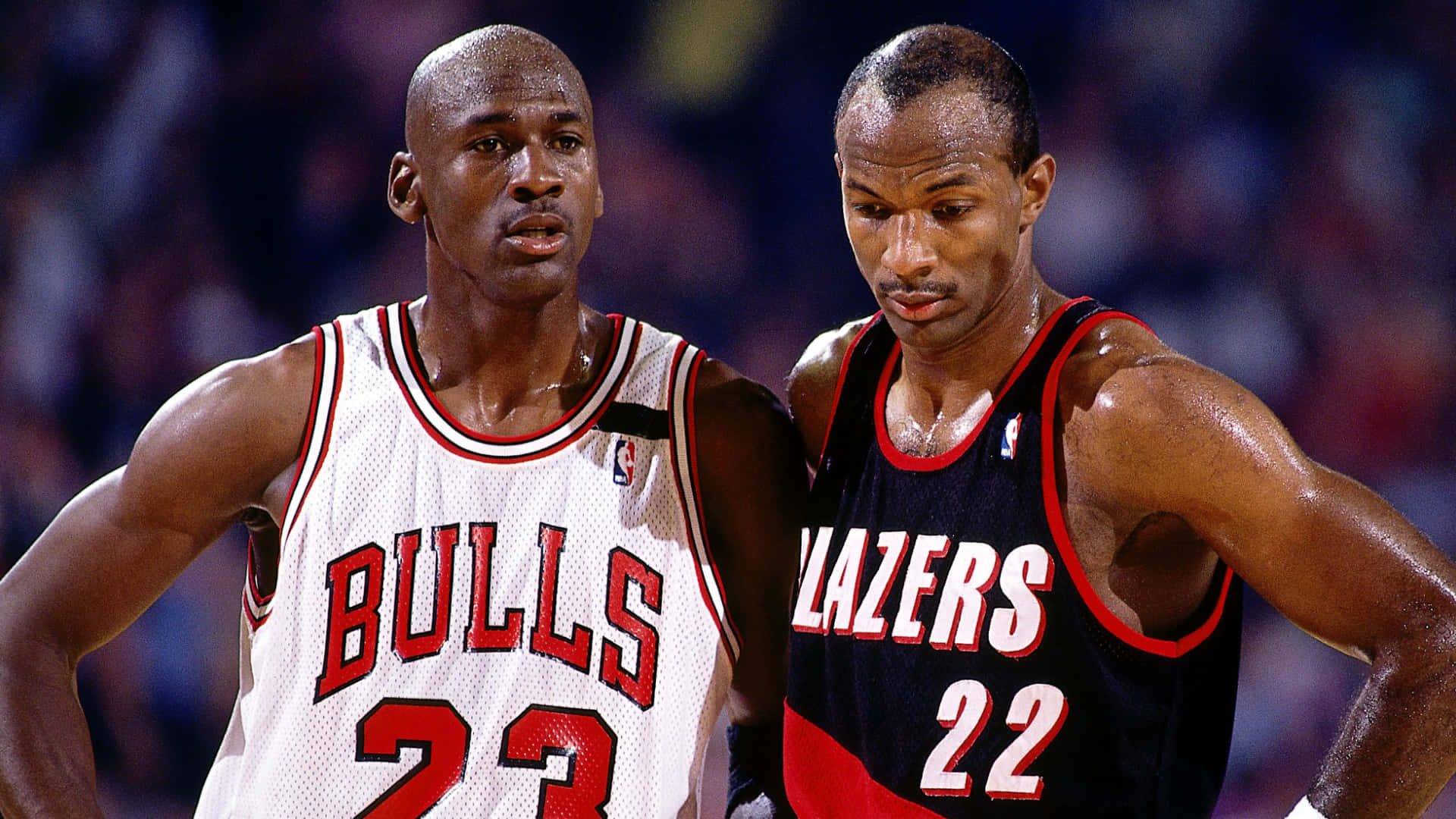 籃球 桌布 壁紙 — 兩位專注於比賽中的激烈NBA球員，一位身穿公牛隊球衣，另一位身穿拓荒者隊球衣，汗水在場館燈光下閃閃發光。