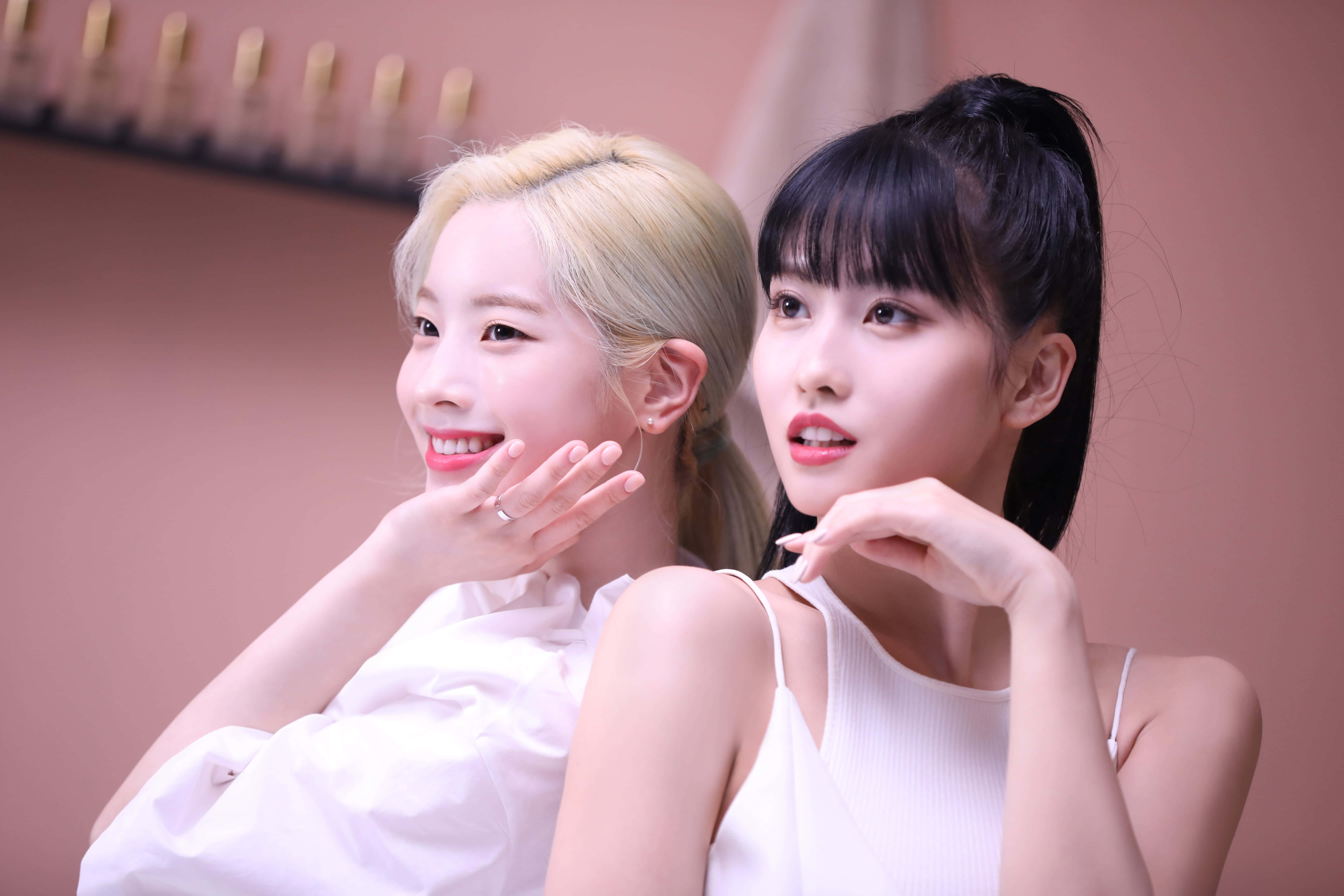 twice 桌布 壁紙 — 兩位年輕女性，一頭金髮一頭黑髮形成對比，在柔和的粉紅色背景前穿著白色上衣俏皮地擺姿勢。