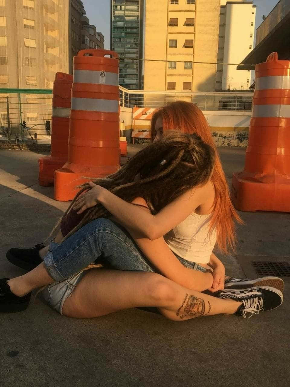 女同志 桌布 壁紙 — 兩位年輕女性，一頭鮮紅色和深色辮子，在陽光明媚的城市人行道上熱情擁抱，周圍是橙色交通錐。