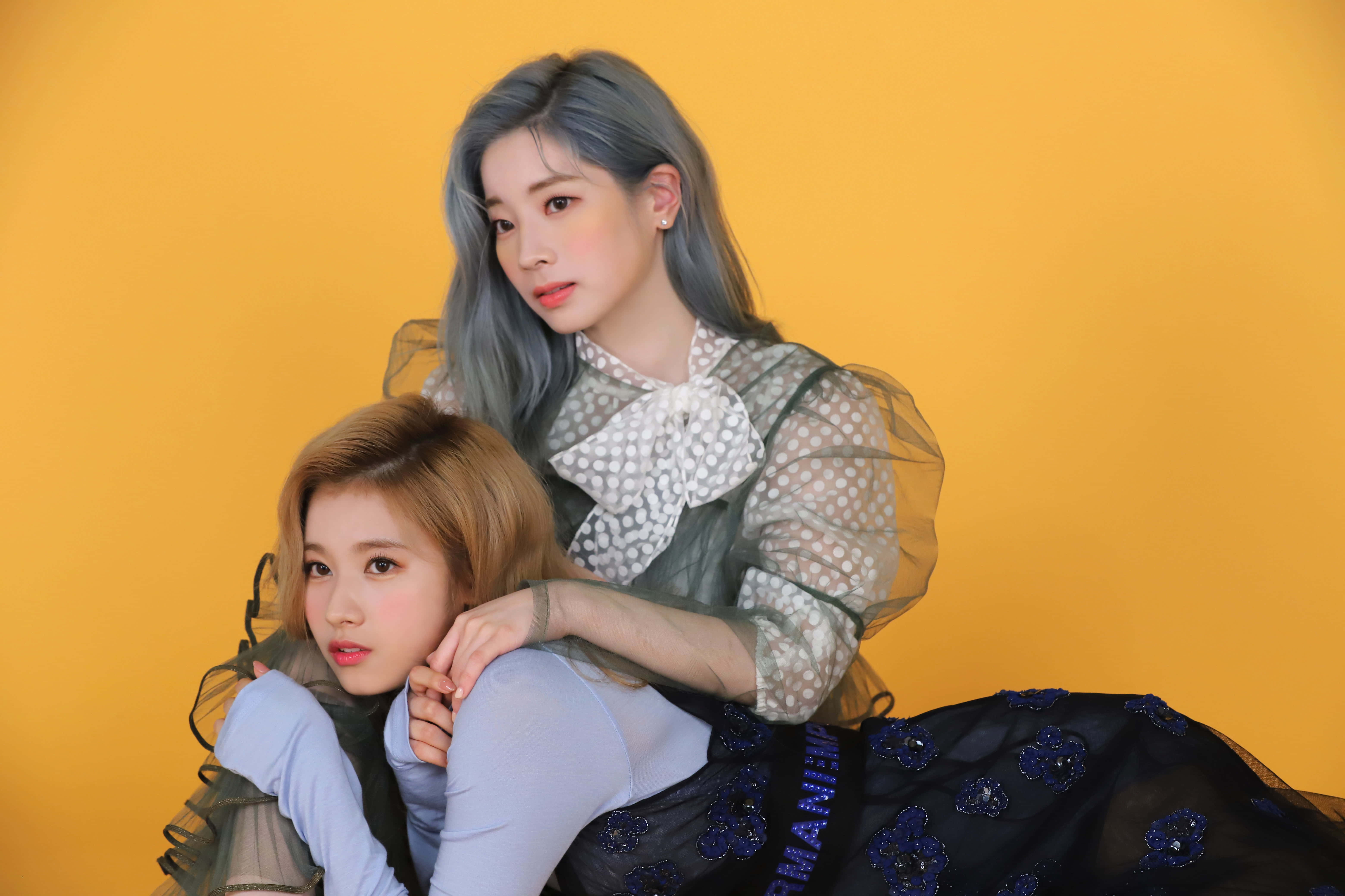 twice 桌布 壁紙 — 兩位年輕女性，頭髮經過造型，在純色溫暖黃色背景前親密擺姿勢，一位穿著圓點襯衫，另一位穿著帶花卉裝飾的藍色連衣裙。