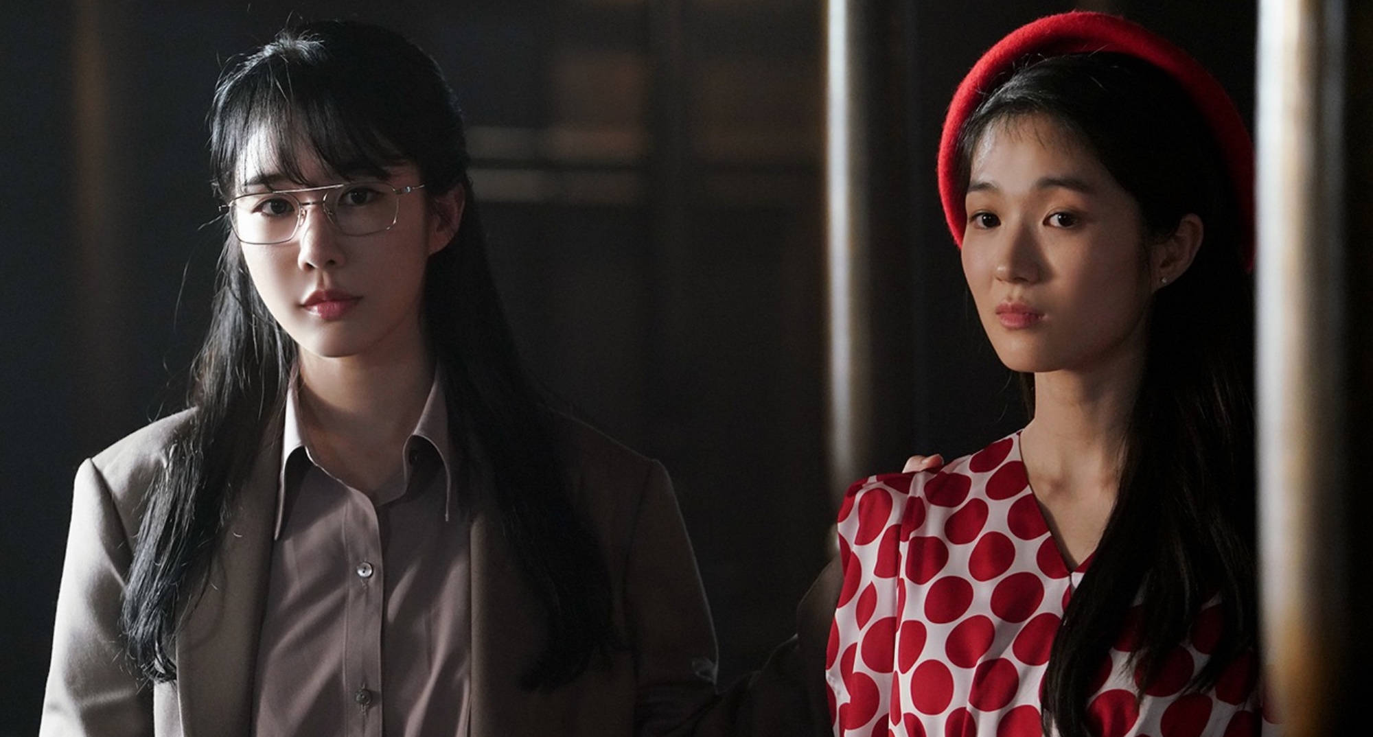 drama 桌布 壁紙 — 兩位年輕女性並排站在燈光昏暗的房間裡，一位戴眼鏡穿米色西裝外套，另一位穿紅色圓點連衣裙和紅色貝雷帽，兩人都表情嚴肅。