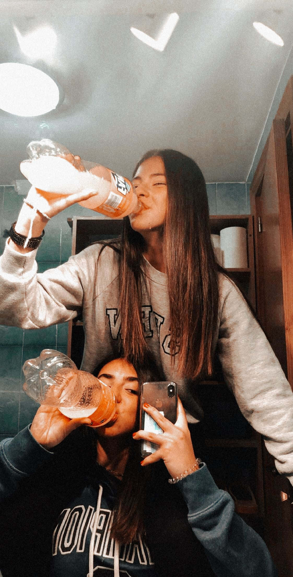 Best Friend Aesthetic 桌布 壁紙 — 兩位年輕女性在浴室鏡前自拍，一邊從塑膠瓶喝水，一邊在明亮的吊燈下。