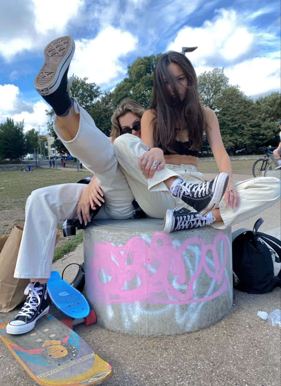friend 桌布 壁紙 — 兩位年輕女性在陽光明媚的公園裡，隨意地躺在塗鴉覆蓋的混凝土柱上，附近有滑板。