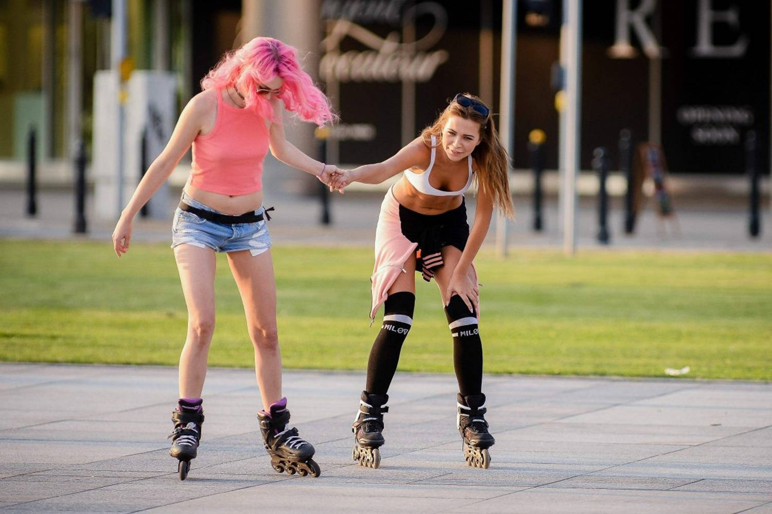 skating 桌布 壁紙 — 兩位年輕女性手牽手在城市人行道上溜冰，身後有草地和商店門面。