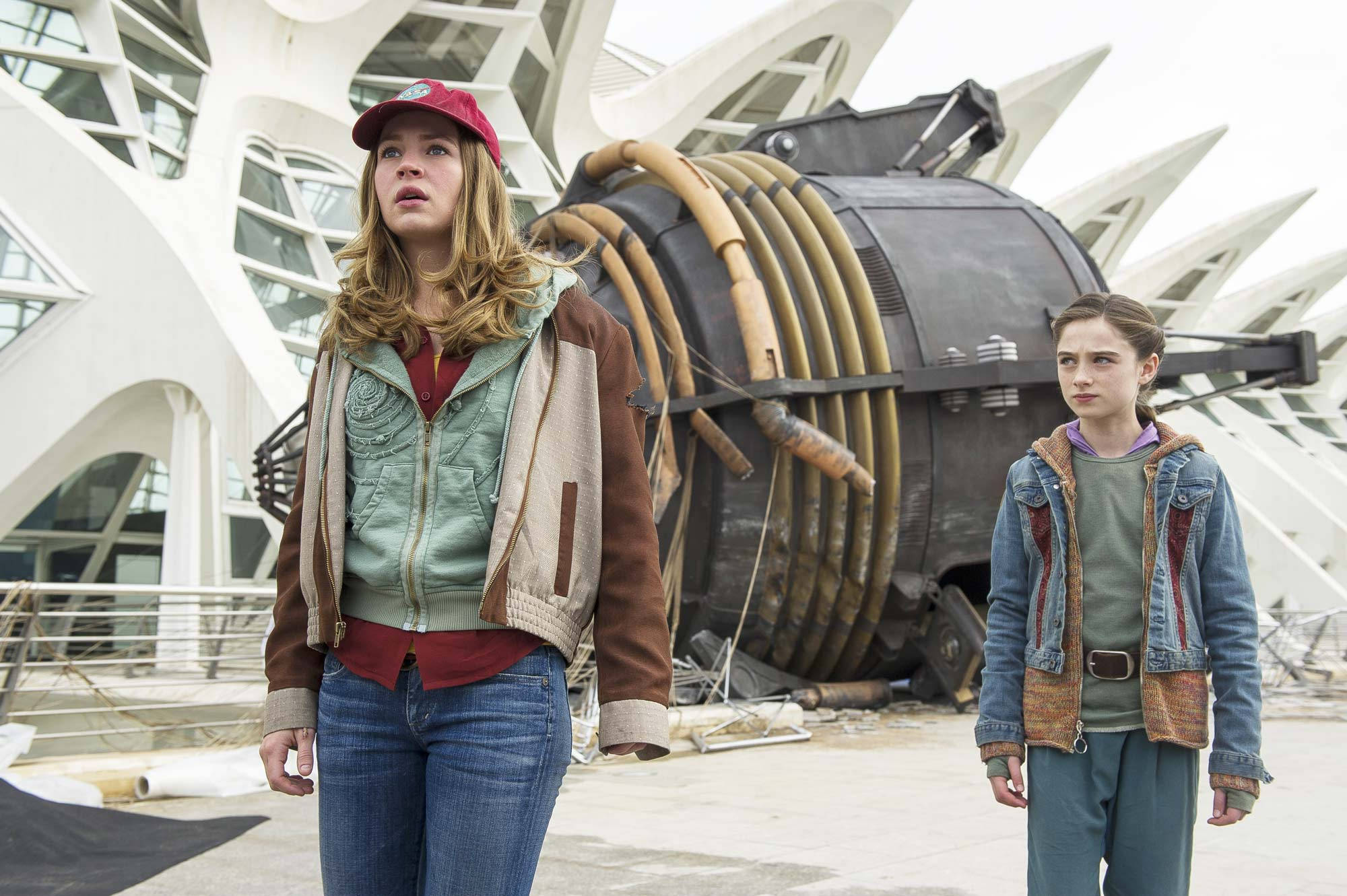 Tomorrowland Movie 桌布 壁紙 — 兩位年輕女性敬畏地站在一艘巨大的未來主義太空船殘骸前，該殘骸撞上了一棟白色現代主義建築。