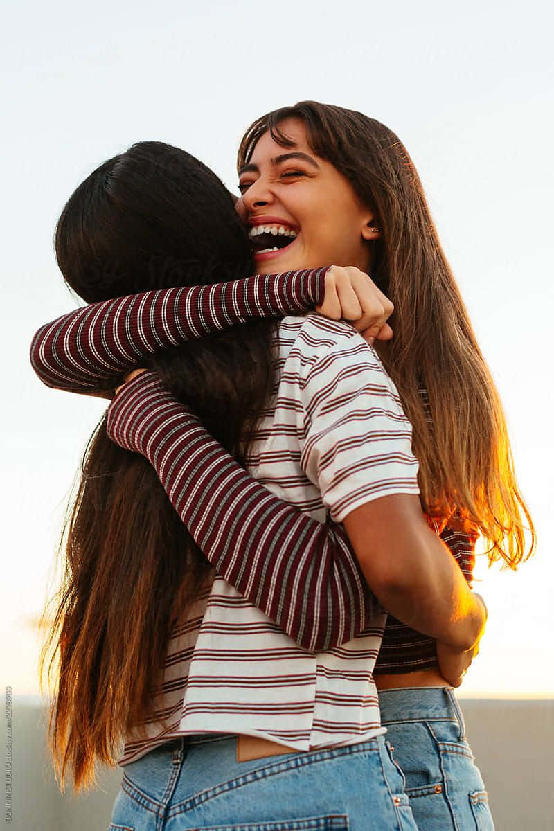 Hugging 桌布 壁紙 — 兩位年輕女性熱情擁抱，金色夕陽灑在她們歡樂的笑聲和條紋上衣上。