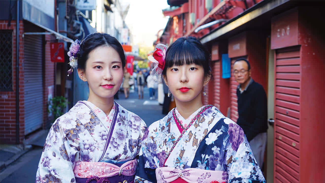 Tokyo Japan 桌布 壁紙 — 兩位年輕女性穿著鮮豔的花卉和服，站在由紅色木建築構成的東京狹窄小巷中。