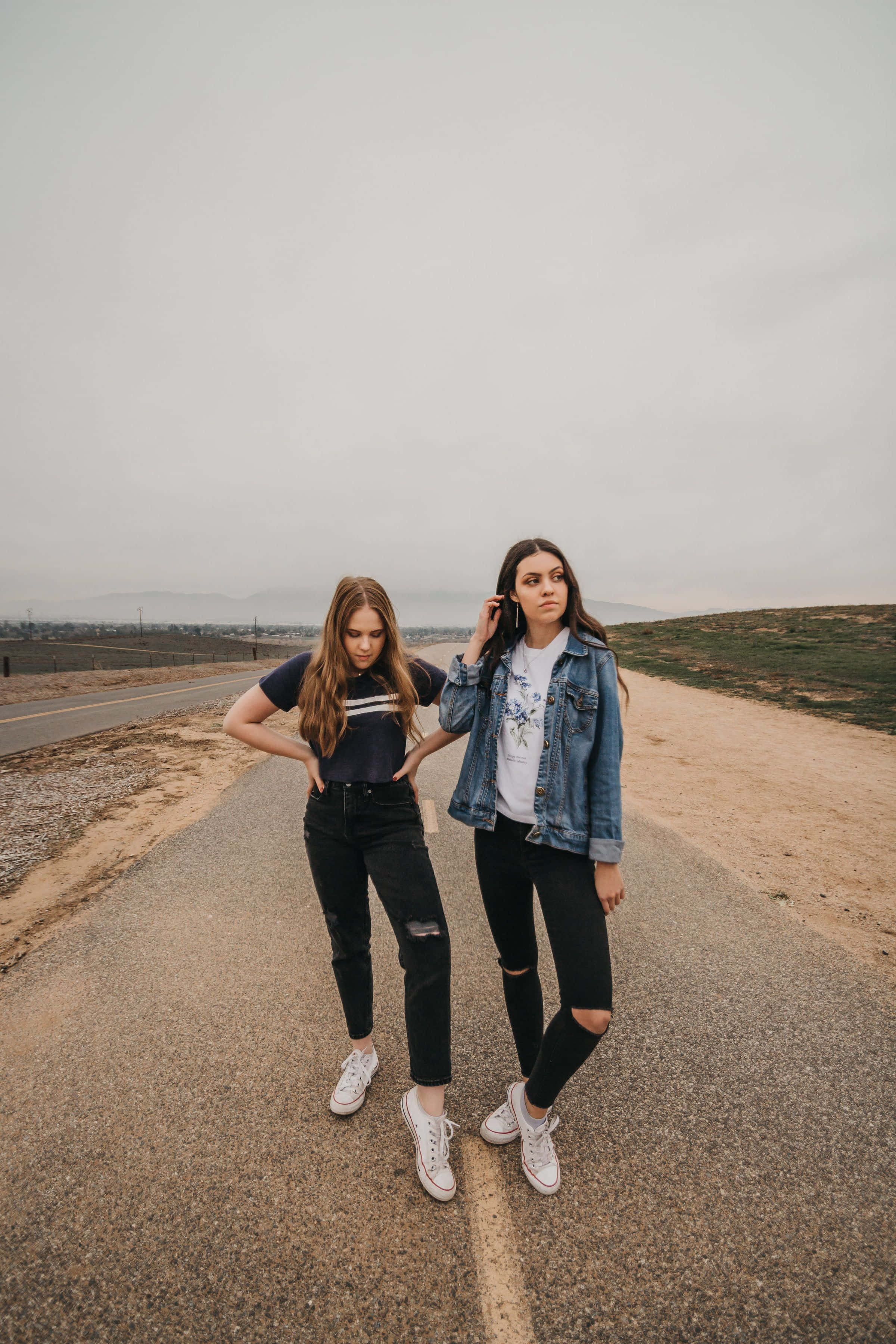 Best Friend Aesthetic 桌布 壁紙 — 兩位年輕女性自信地站在一條荒涼的道路上，天空多雲，遠處有山丘。