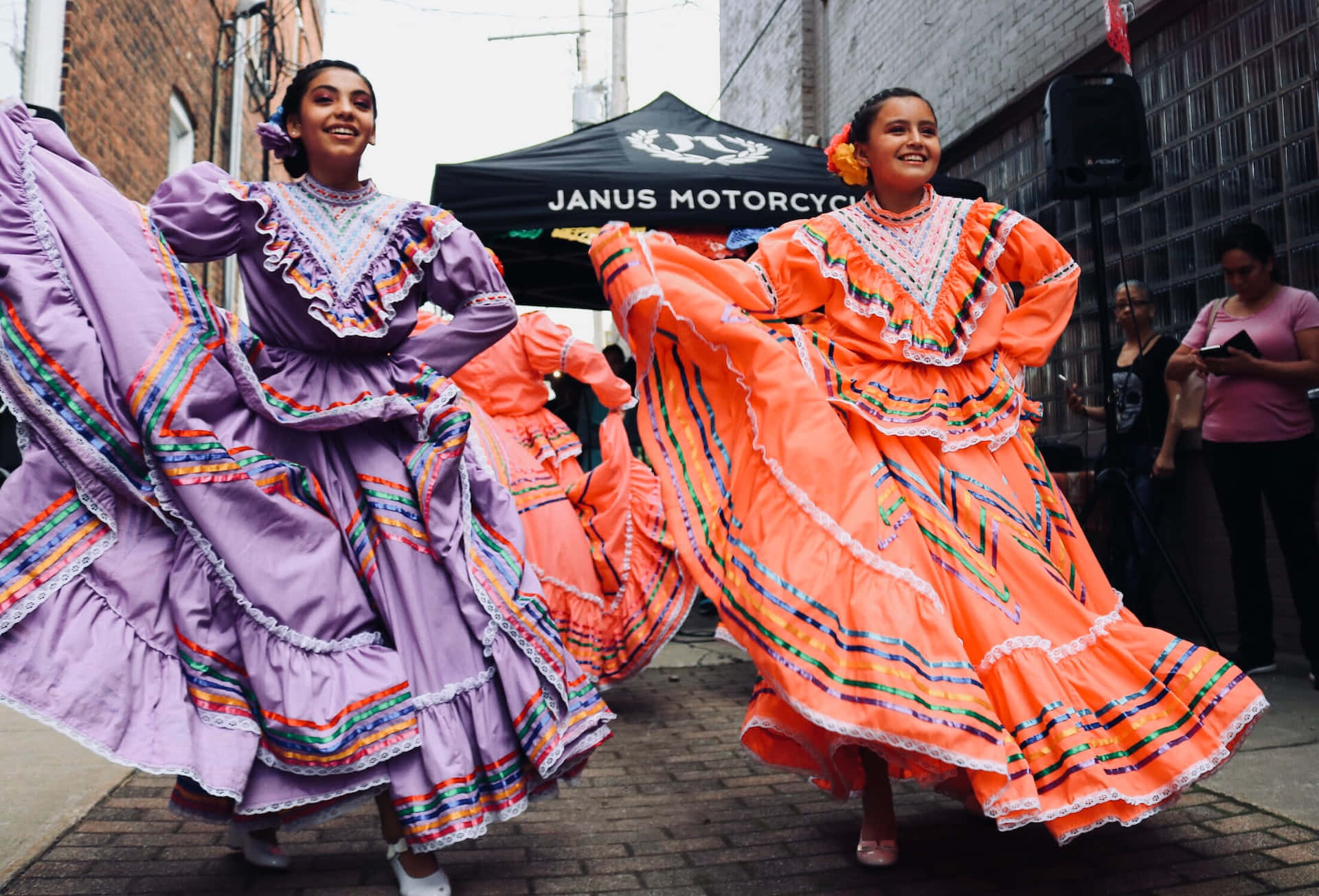 Hispanic 桌布 壁紙 — 兩位年輕女性身穿鮮豔的傳統服飾，在熱鬧的文化慶典期間，在磚街上歡快地旋轉。