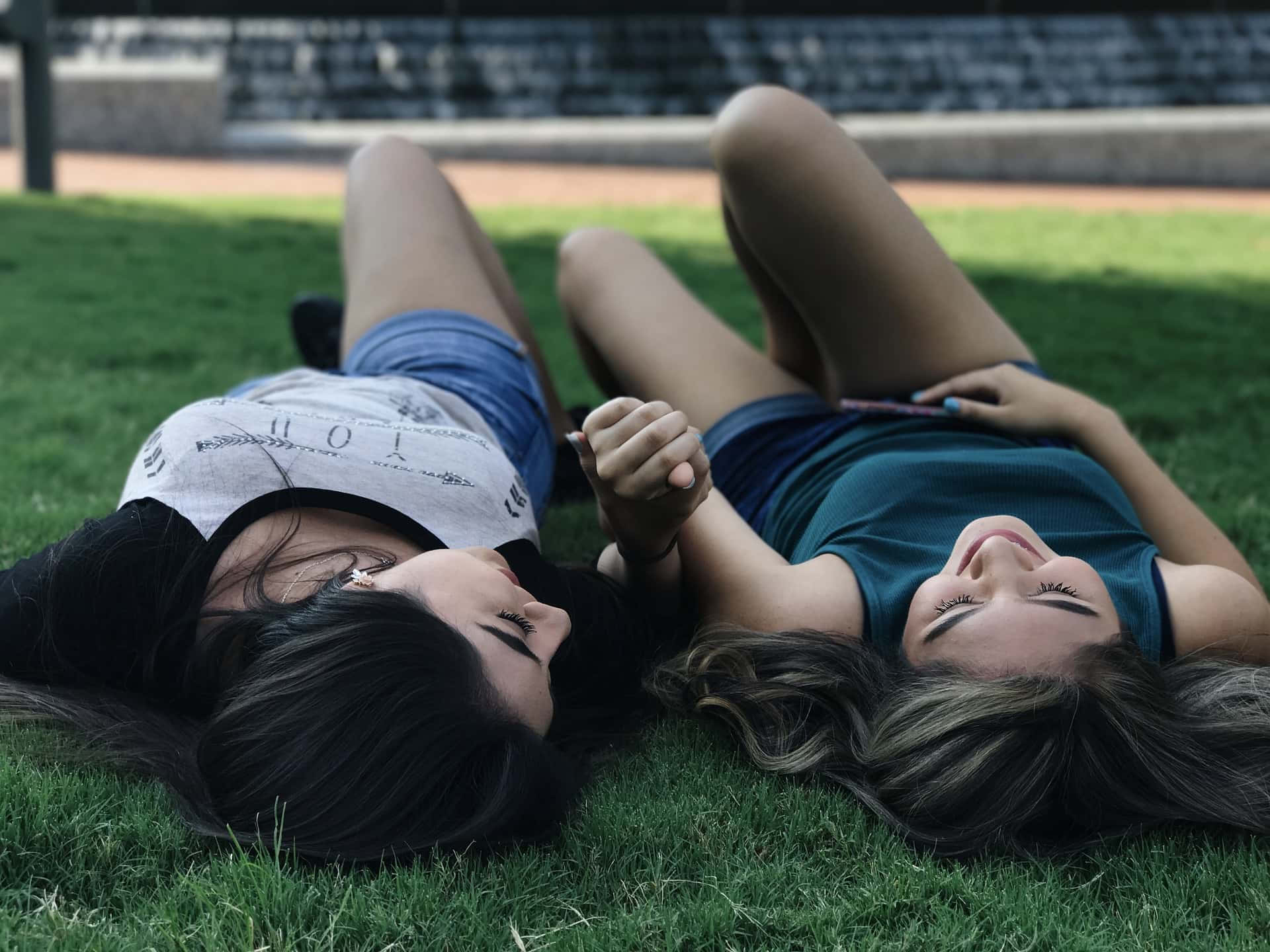 Grass 桌布 壁紙 — 兩位年輕女性躺在草地上，手牽手，在明亮的陽光下互相微笑。