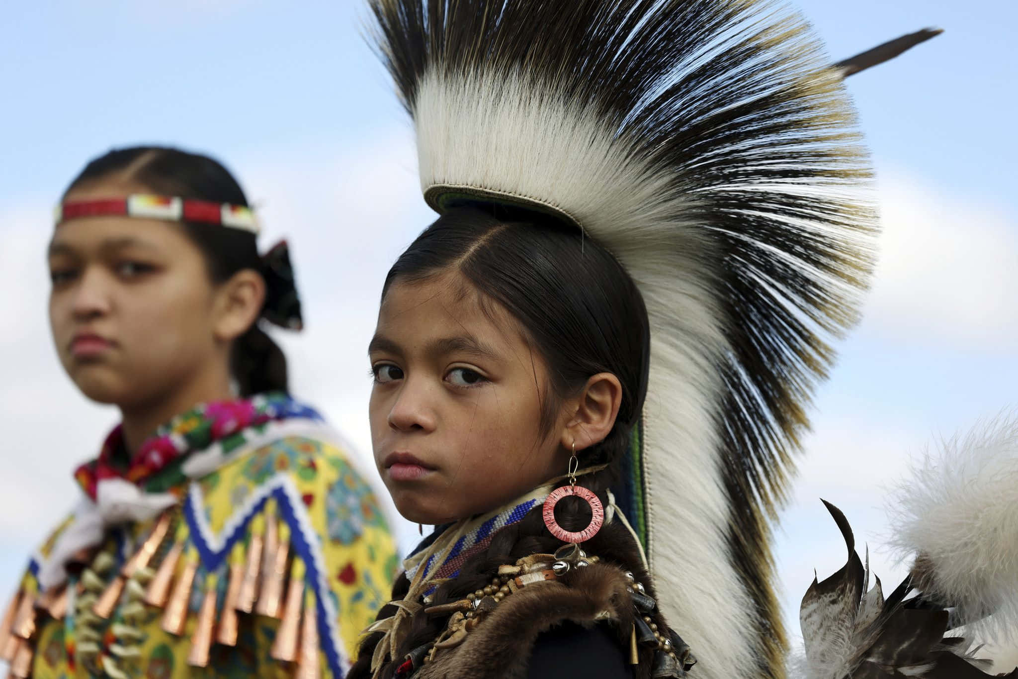 Native American 桌布 壁紙 — 兩位年輕的原住民女孩，戴著傳統羽毛頭飾和串珠禮服，在明亮的天空下莊嚴地凝視。