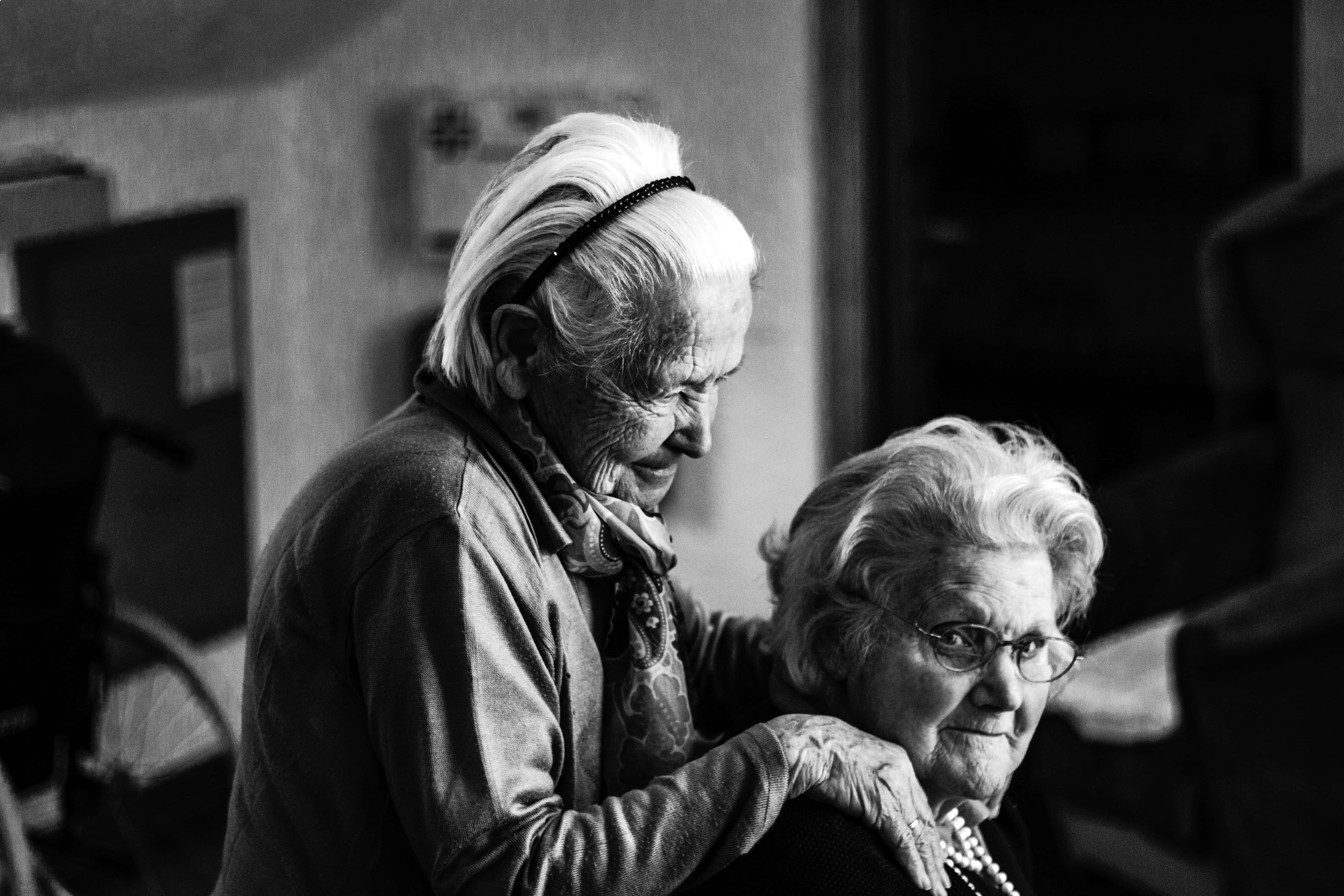 Woman 桌布 壁紙 — 兩位年長女性在燈光昏暗的房間裡分享溫馨時刻，其中一位輕輕將手放在另一位肩上。