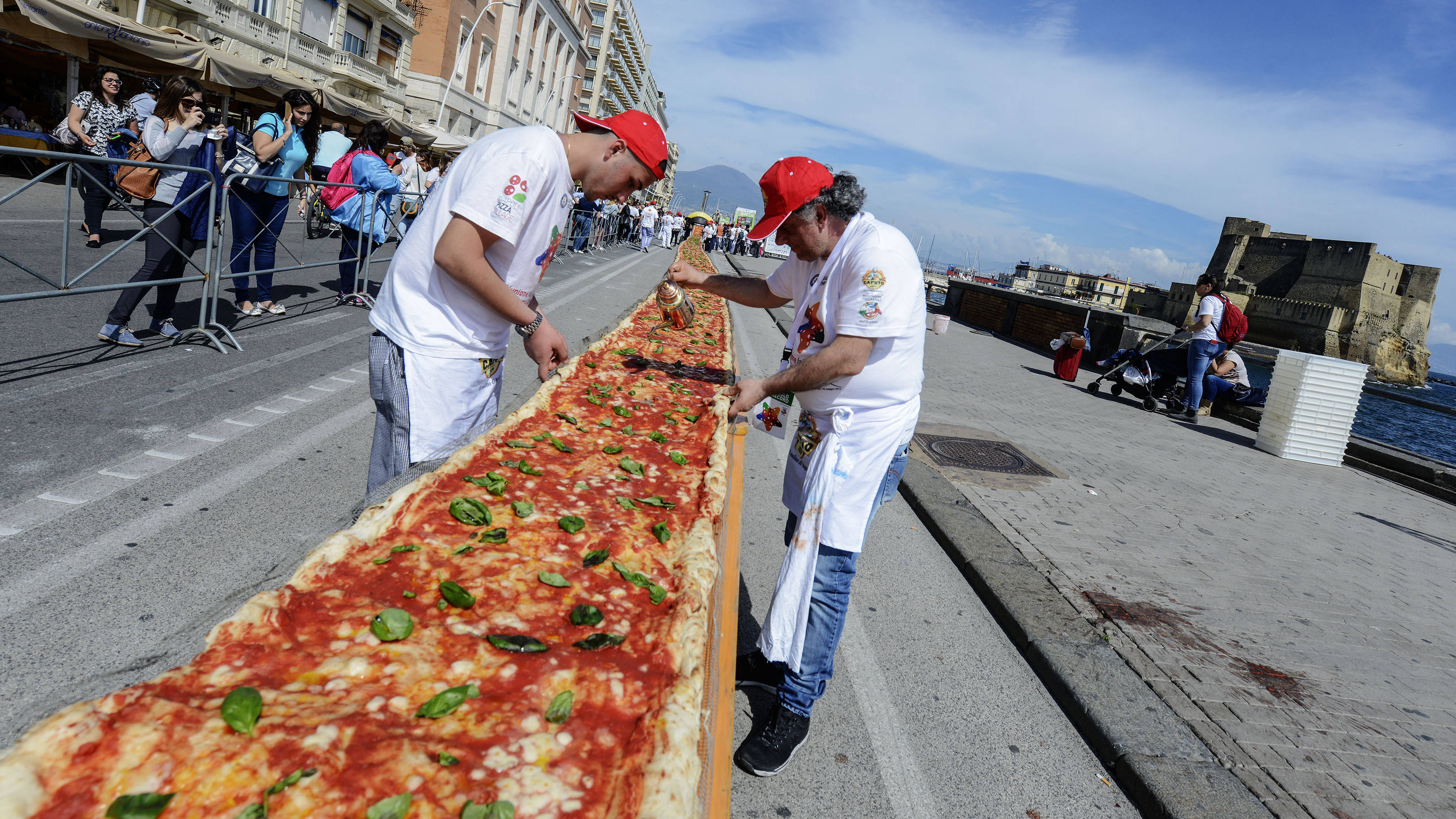Pizza 桌布 壁紙 — 兩位廚師在海濱長廊上切開一塊巨大的長方形披薩，觀眾在明亮的藍天下觀看。