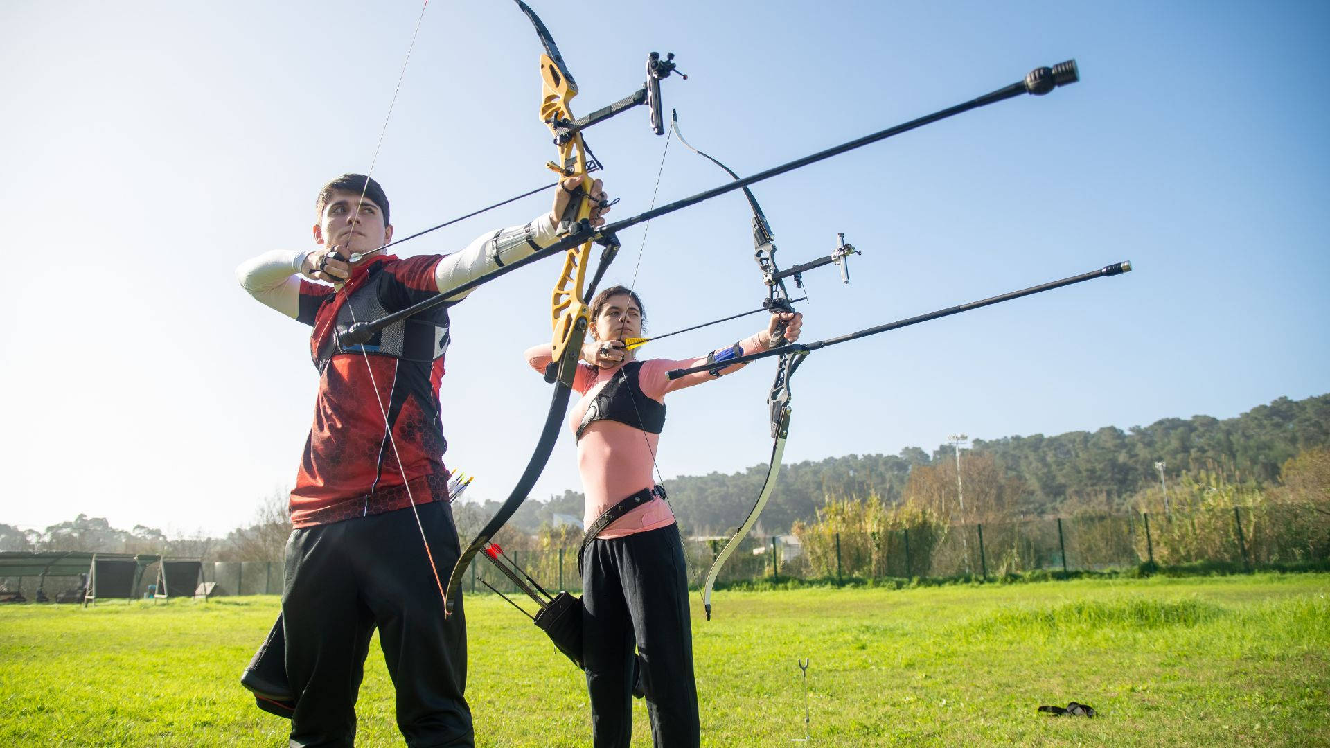 Archery 桌布 壁紙 — 兩位弓箭手以專注姿態拉弓，身處陽光普照的草地，背景有樹木。