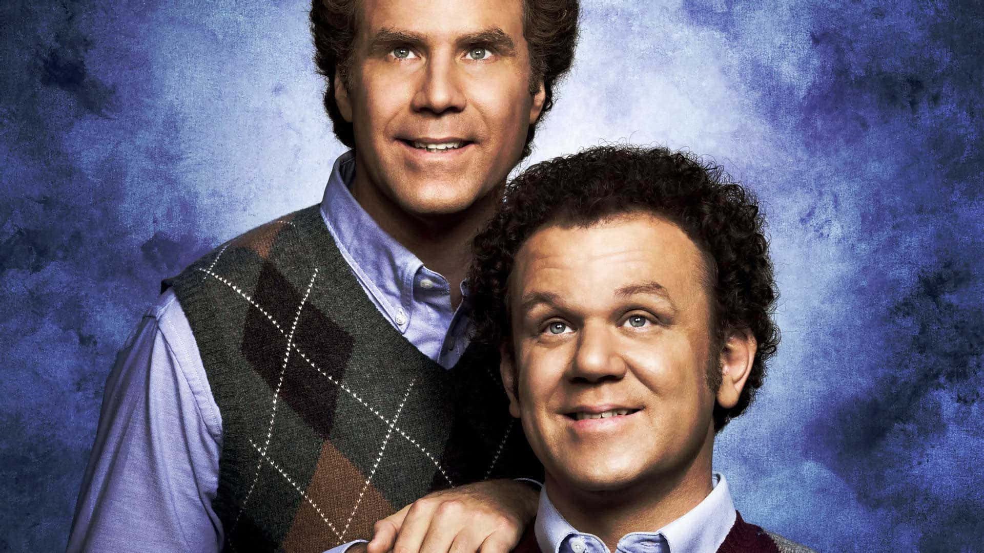 John C. Reilly 桌布 壁紙 — 兩位微笑男士穿著菱形花紋背心，在溫暖燈光下以紋理藍色背景前擺姿勢。