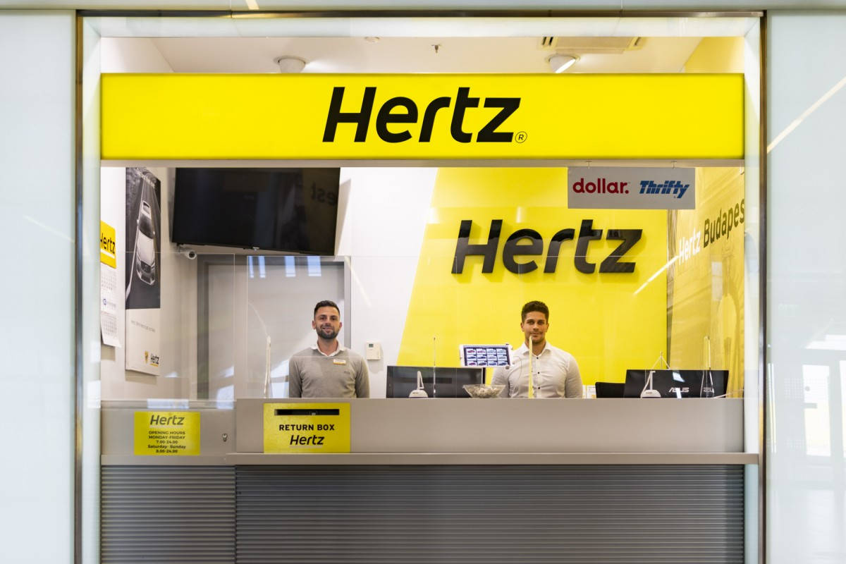Promotion 桌布 壁紙 — 兩位微笑的Hertz員工站在現代租車辦公室內一個亮黃色品牌櫃檯後面。