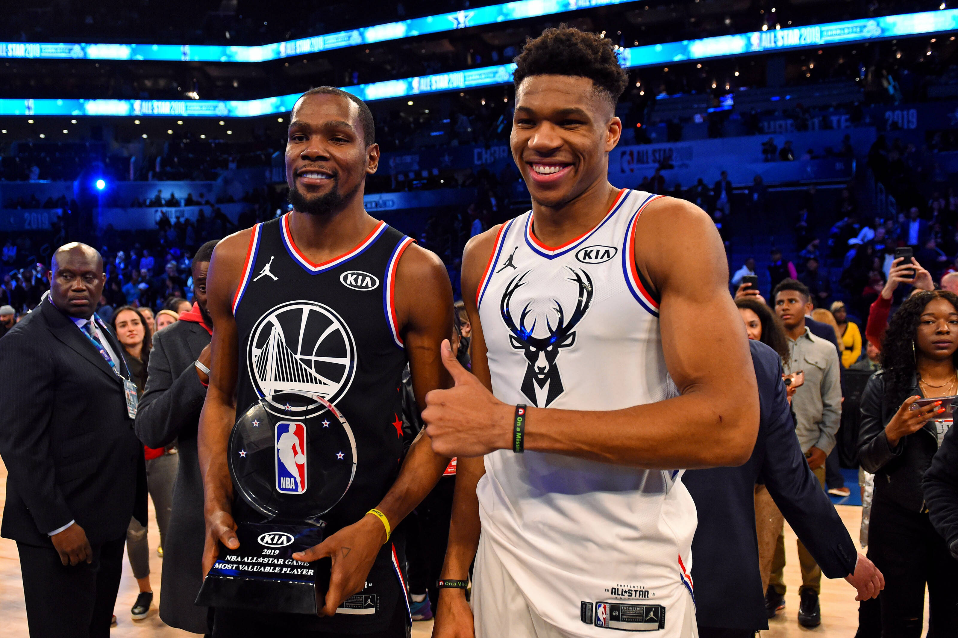 Giannis 桌布 壁紙 — 兩位微笑的NBA球員在燈光明亮的球場上與獎盃合影，周圍是歡呼的球迷。