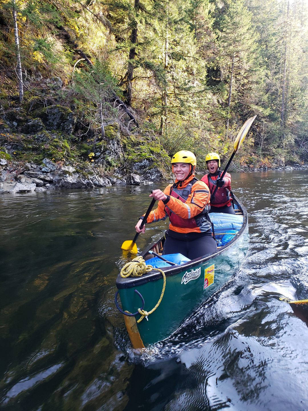 Rowing 桌布 壁紙 — 兩位微笑的冒險家划著綠色獨木舟穿過森林河流，河中有長滿苔蘚的岩石和高大的樹木。