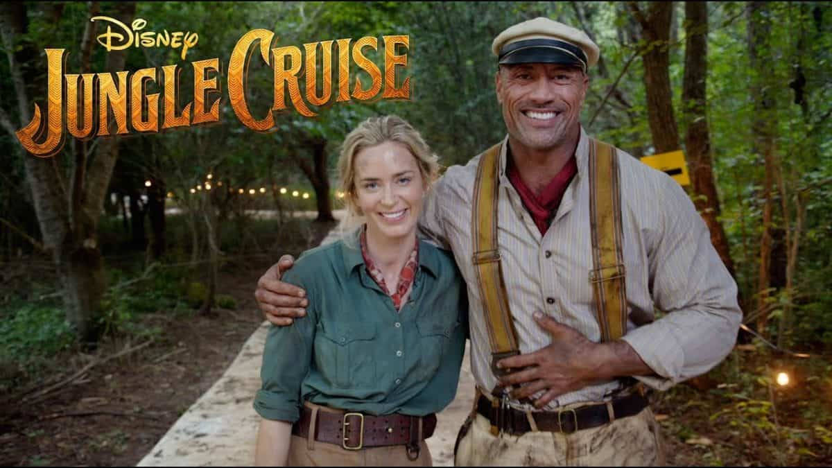 Jungle Cruise 桌布 壁紙 — 兩位微笑的冒險家穿著叢林探險服裝，站在一條被茂密綠樹環繞的小徑上，背景中溫暖的串燈閃爍著光芒。