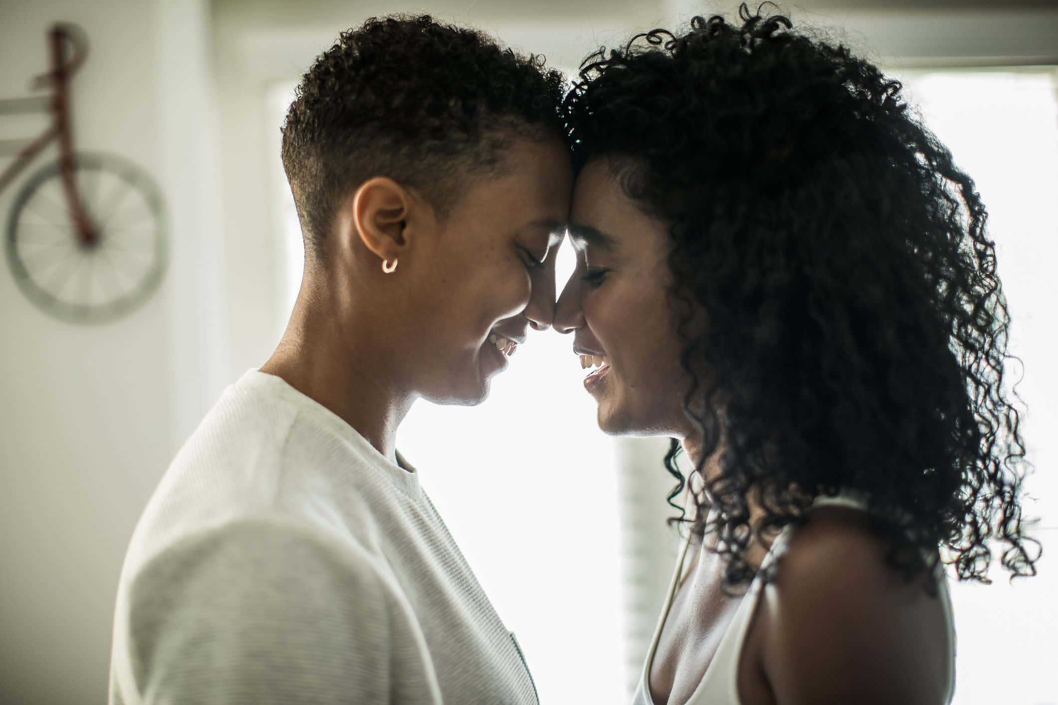 Black Couple 桌布 壁紙 — 兩位微笑的女性，髮質對比鮮明，在柔和光線的室內空間親密地額頭相觸。