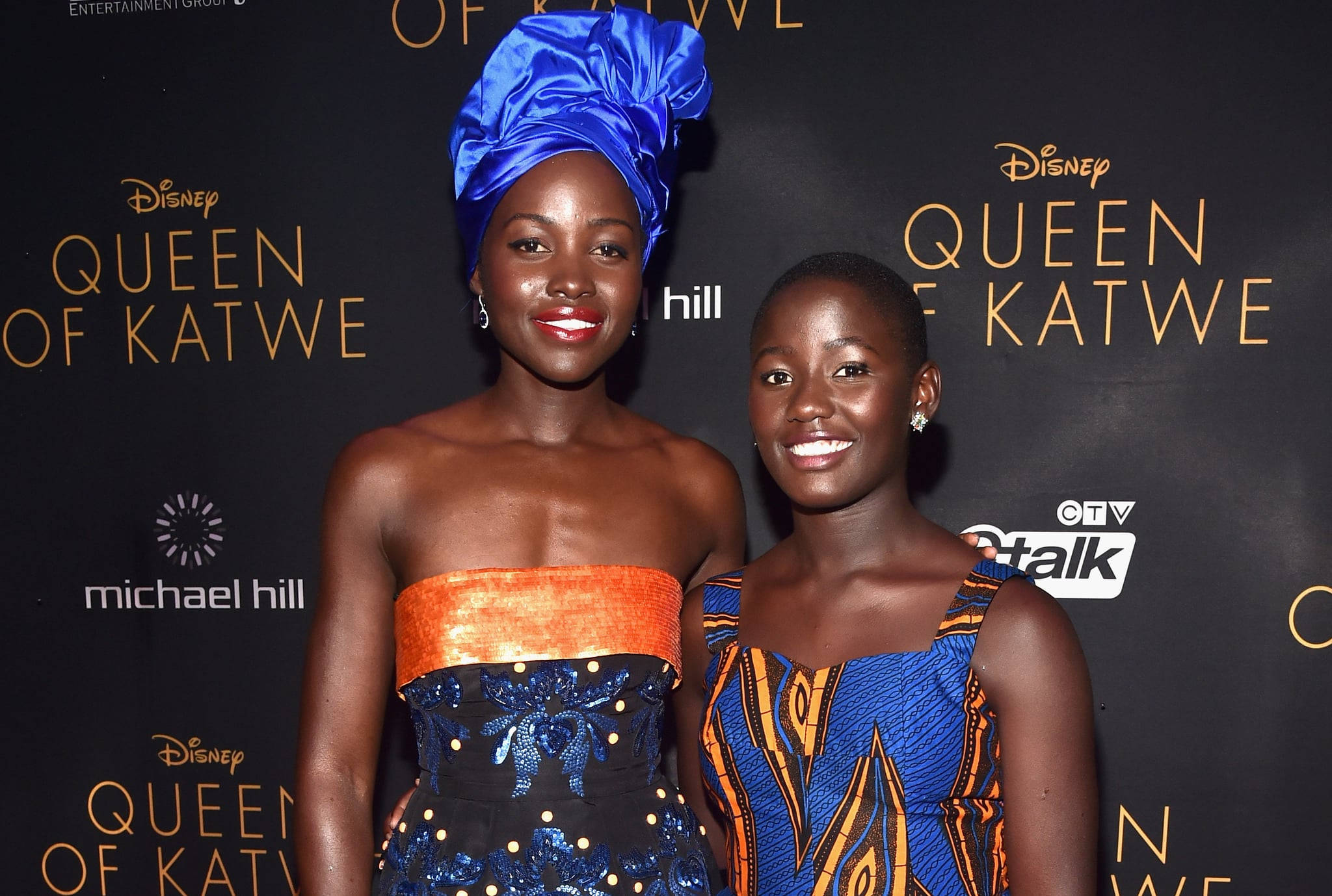 queen of katwe 桌布 壁紙 — 兩位微笑的女性在《Queen of Katwe》首映會上，穿著鮮豔的藍色和橙色傳統服飾，在品牌背景前擺姿勢。