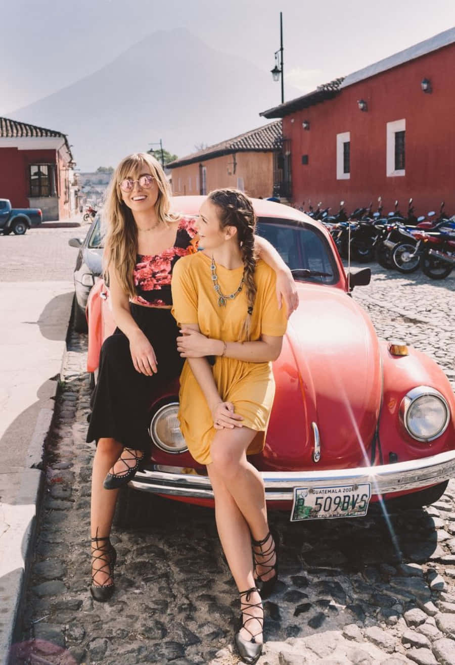 Friends 桌布 壁紙 — 兩位微笑的女性在一輛停泊在鵝卵石街道上的復古紅色福斯金龜車上擺姿勢，背景有山。